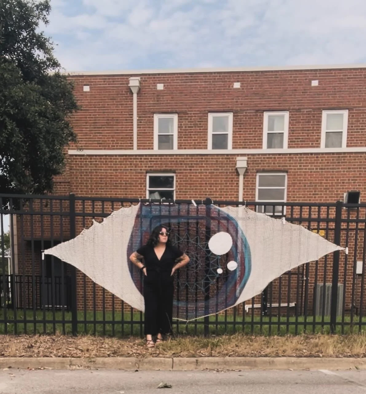 A woman with dark curly hair, wearing sunglasses and black clothing, standing in front of a decorative fence with a mural of an eye behind her.