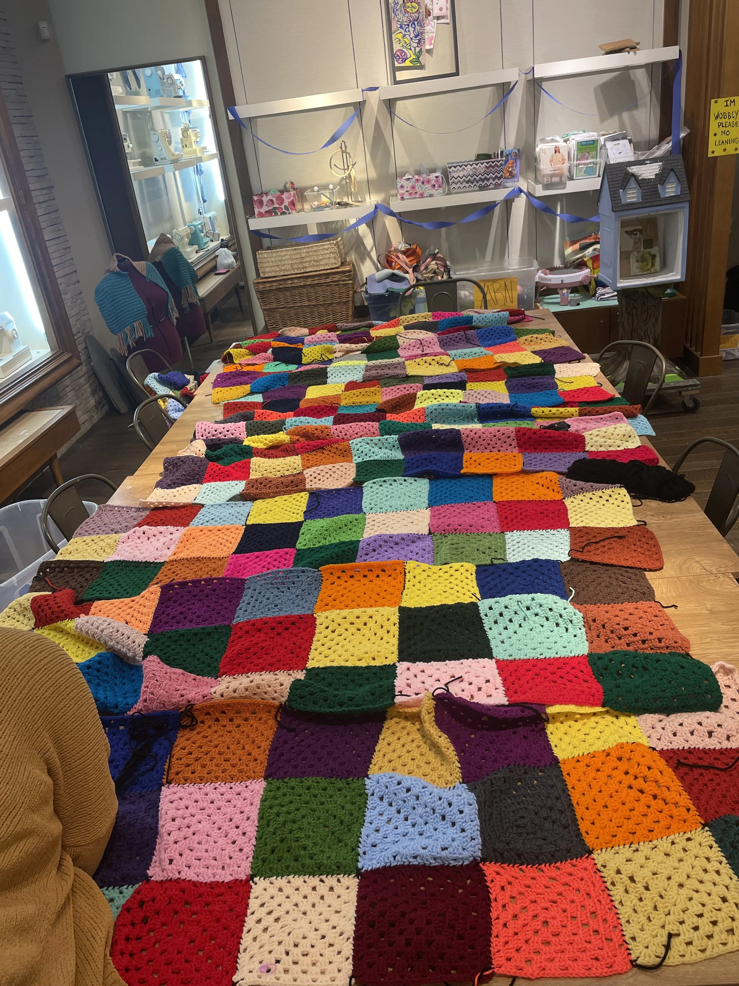 Colorful crochet granny squares blanket displayed on a large wooden table in a craft room, with shelves and decorations in the background.