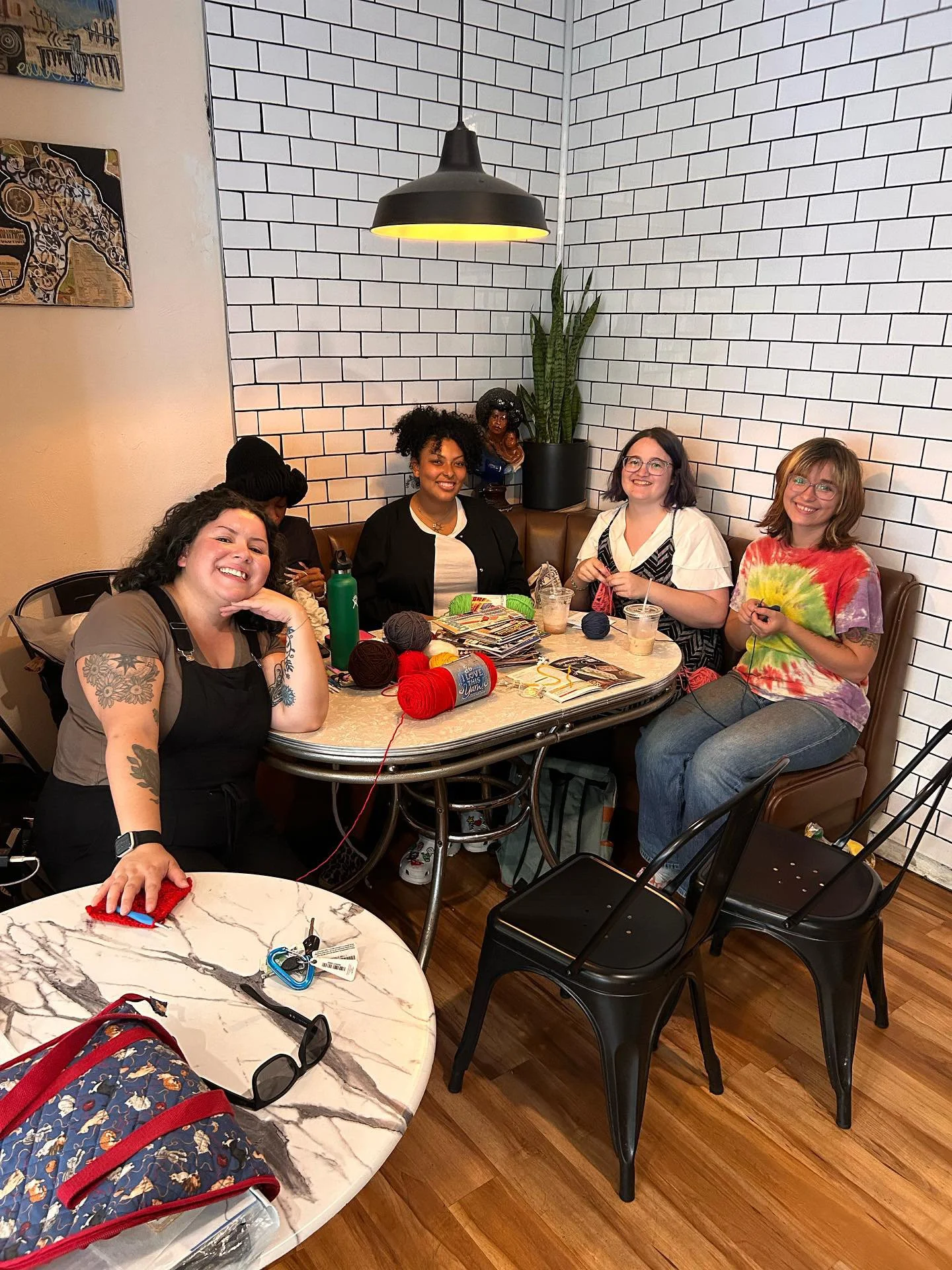 Four women sitting around a table in a cafe, knitting and crocheting, with yarn and crafting supplies on the table.