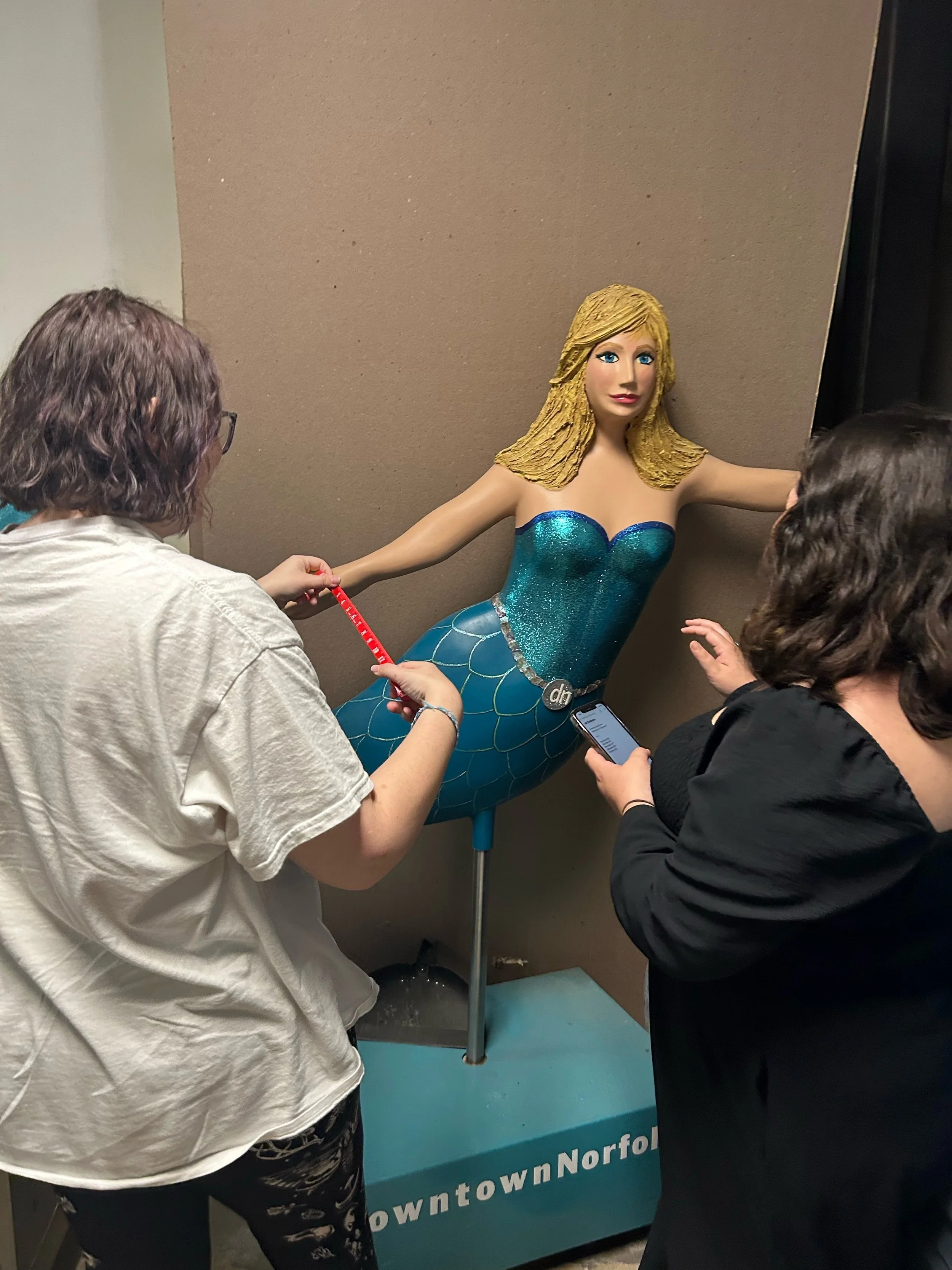 A mermaid sculpture with blonde hair, a blue shimmering tail, and top, is displayed against a beige wall. Two women are examining the sculpture, one holding a measuring tape and the other looking at her phone.