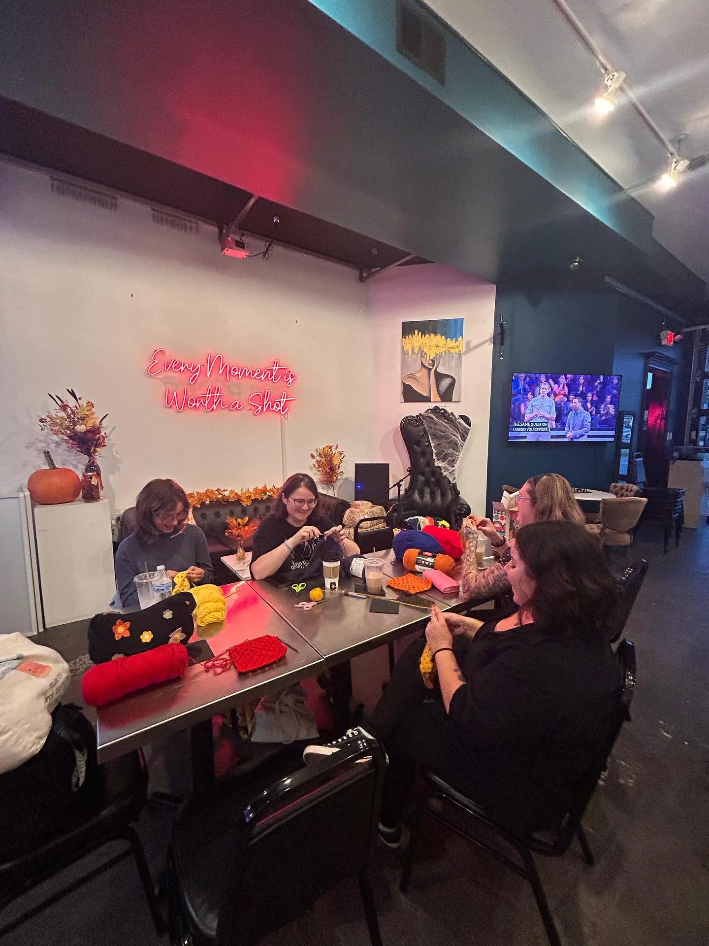 Four women sitting at a table engaging in knitting at a cozy, decorated craft night gathering with a neon sign on the wall that says 'Every Moment is Worth a Shot'. The room features autumnal decorations, paintings, and a television on the wall.