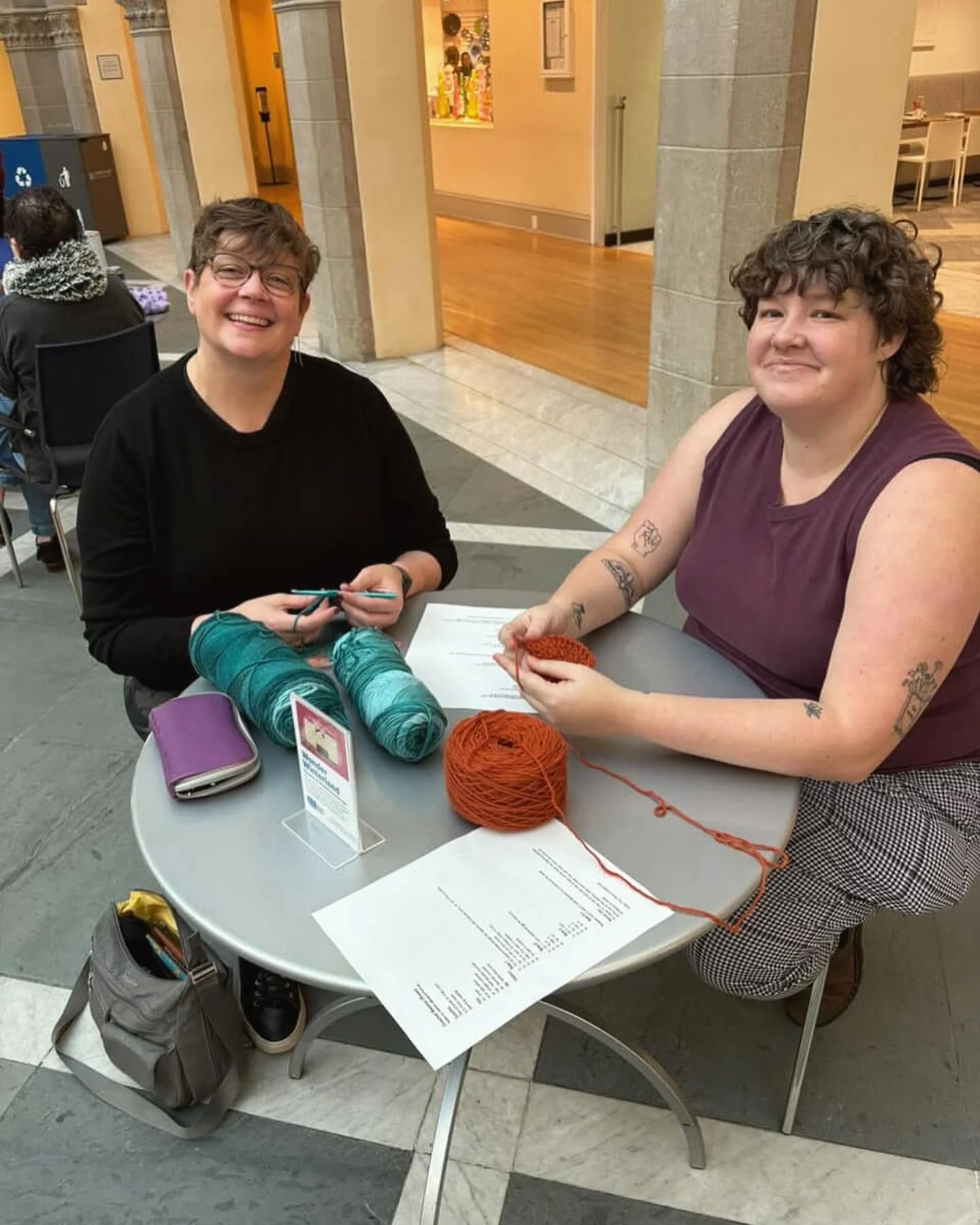 Two women sitting at a table with yarn, knitting needles, and knitting project in a public indoor space, smiling at the camera.