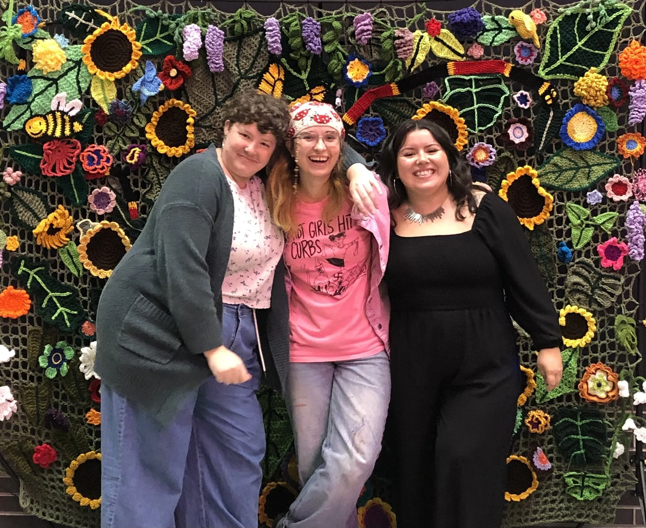 Three women smiling and standing together in front of a colorful flower and leaf crochet wall hanging.