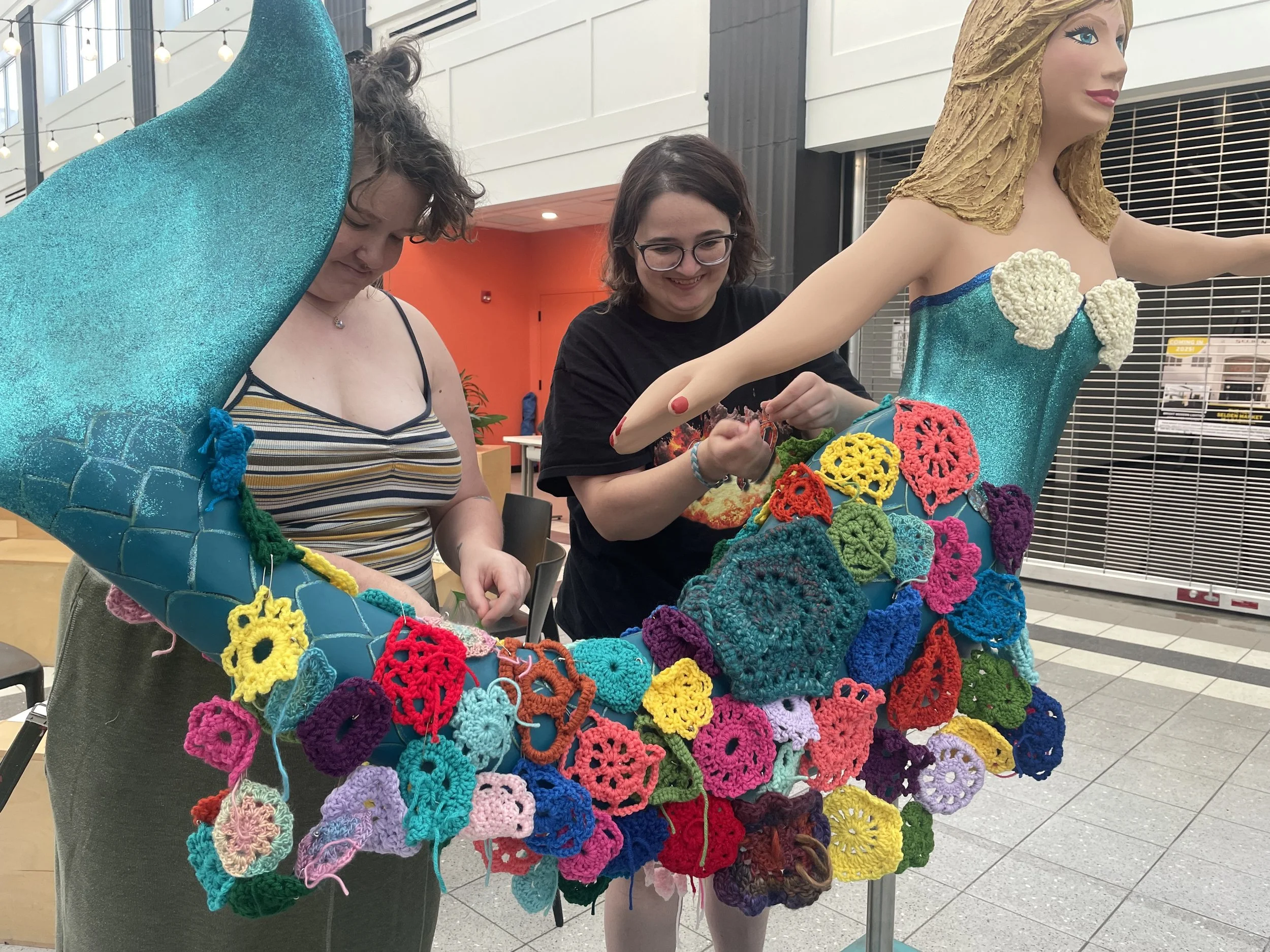 Two women working on a colorful crocheted art installation around a mannequin dressed as a mermaid. They are adding crochet pieces to the installation.