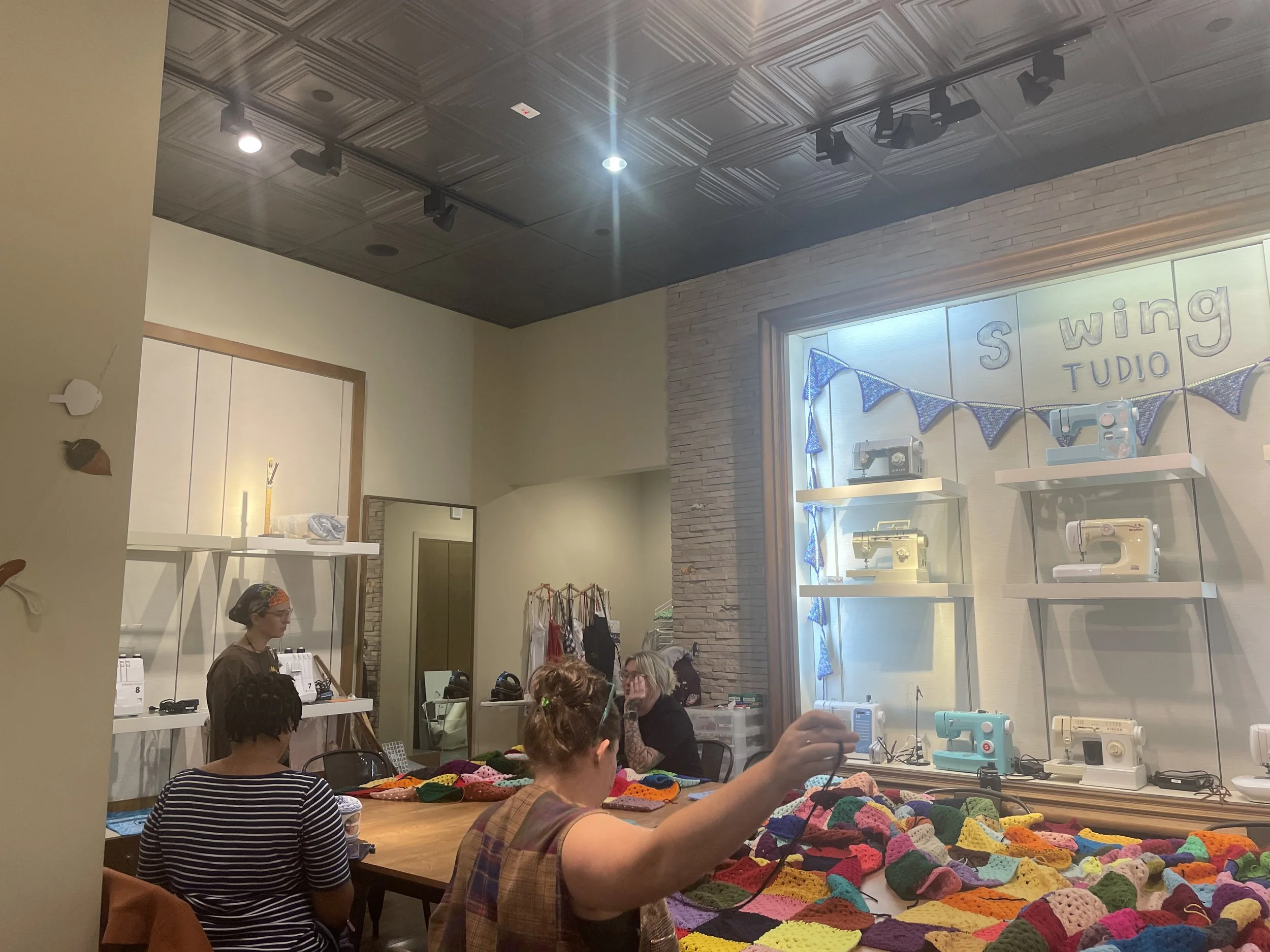 People sitting at a table with colorful crochet items, inside a sewing studio with vintage sewing machines displayed on shelves and a sign that reads 'S Wing Studio'.