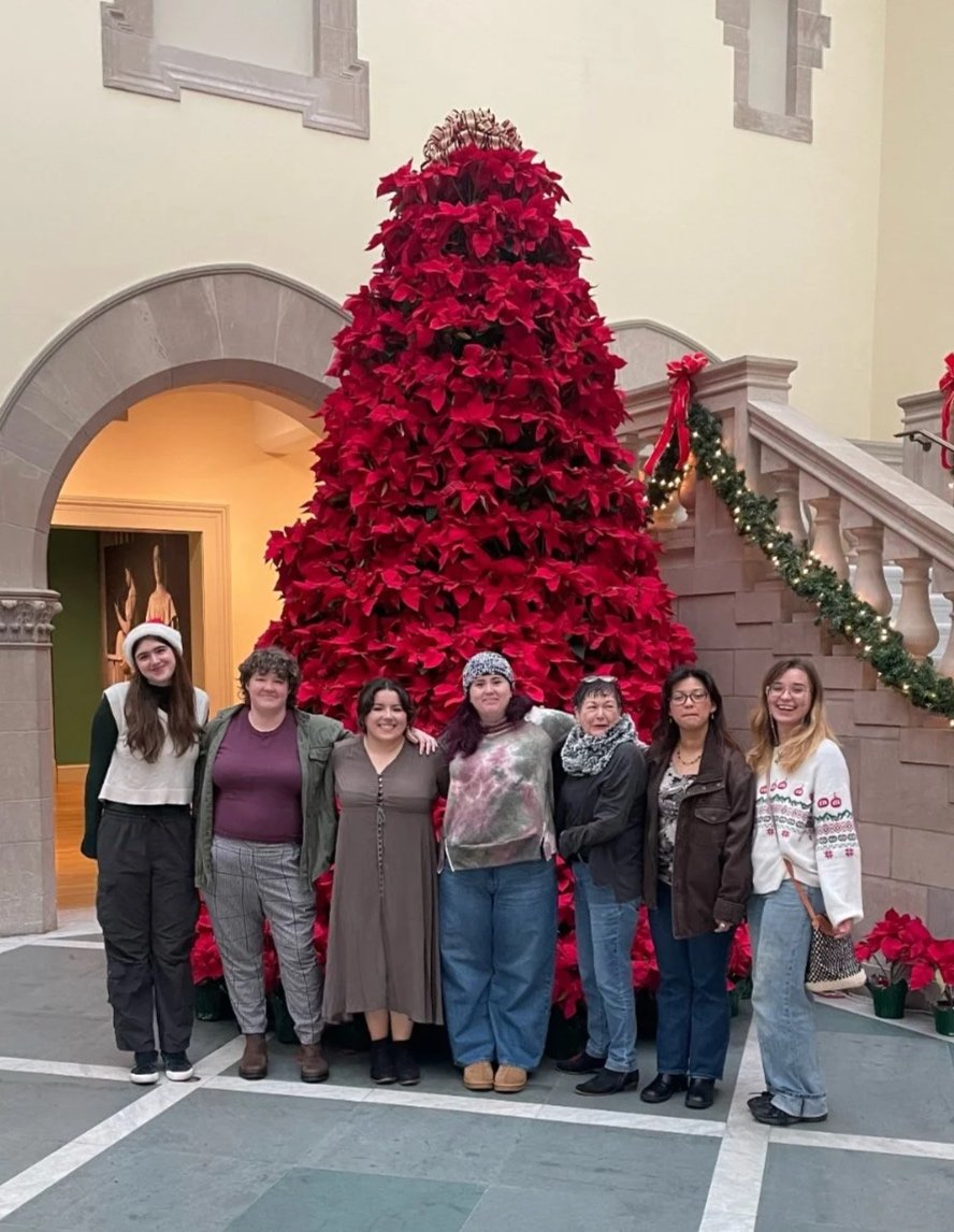 A group of seven women standing in front of a large red poinsettia Christmas tree inside a building decorated with holiday ornaments.