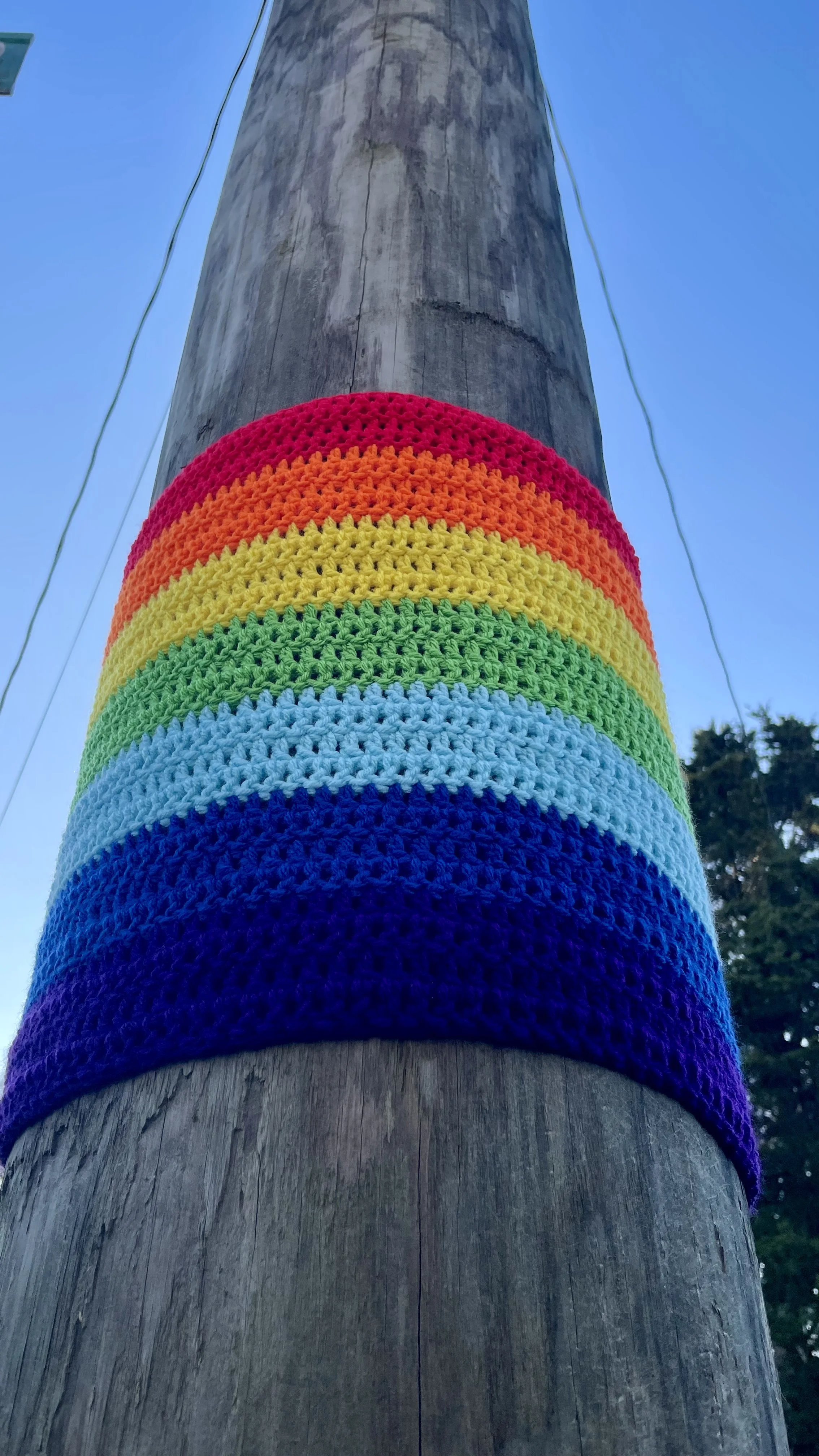 Colorful rainbow crochet pattern wrapped around a wooden utility pole against a blue sky.