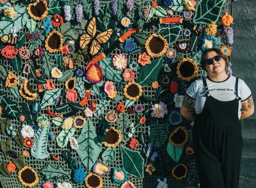 A woman with black hair, wearing sunglasses, a white t-shirt, and black overalls, standing in front of a colorful embroidered floral mural.