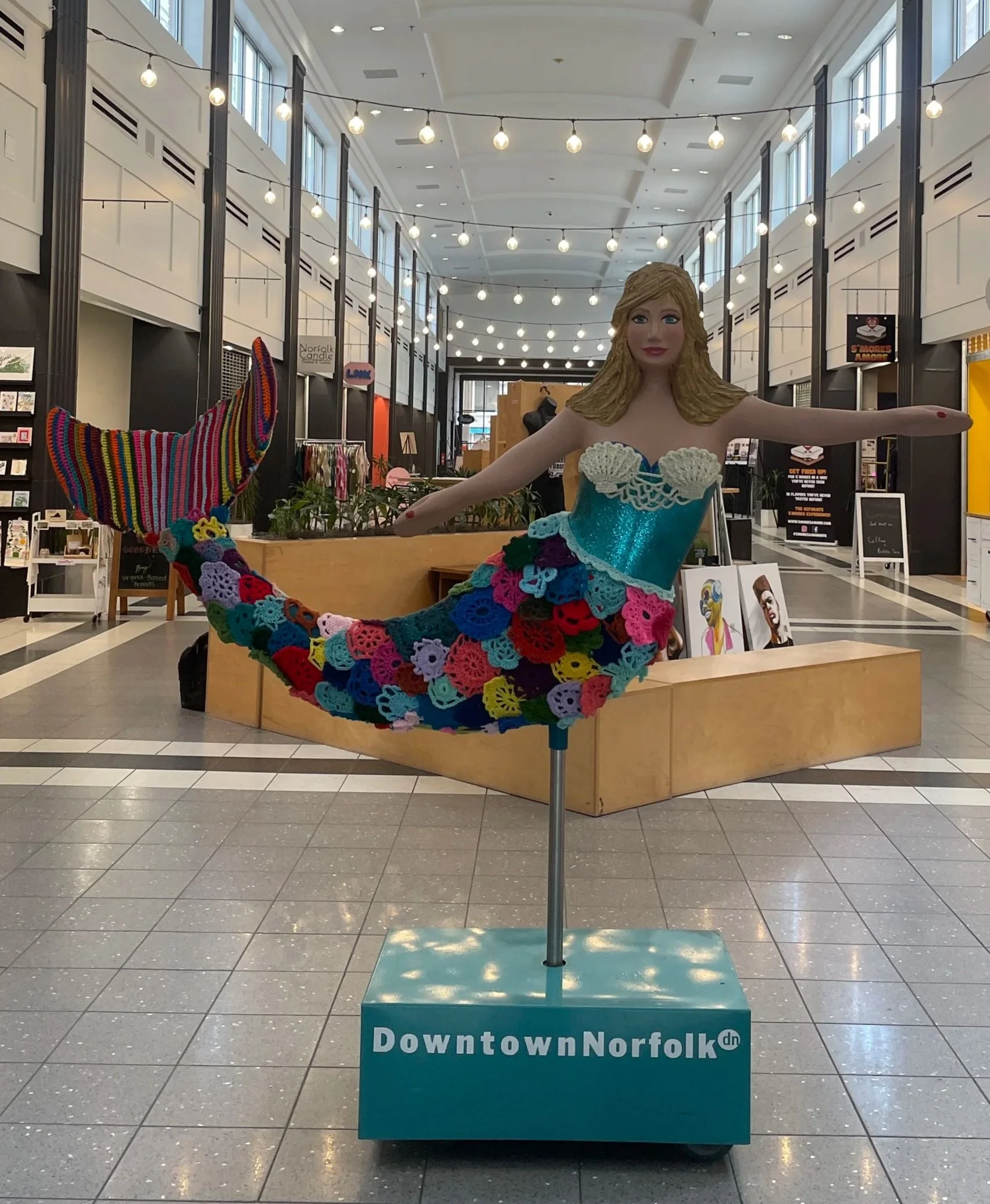 A mannequin with a mermaid tail and top made of colorful crochet with flowers, in a shopping mall with string lights and art displays.
