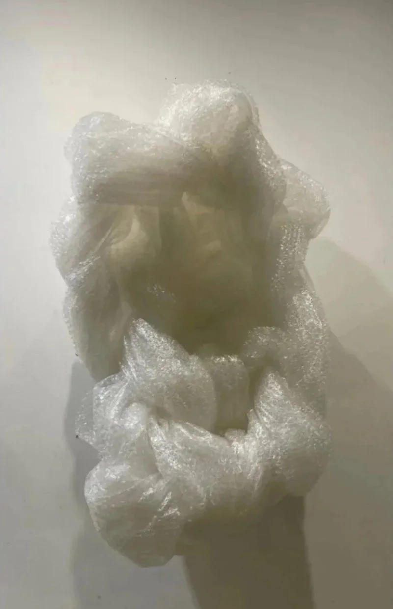 A person wrapped in crumpled clear plastic or cellophane, with only part of their face visible.