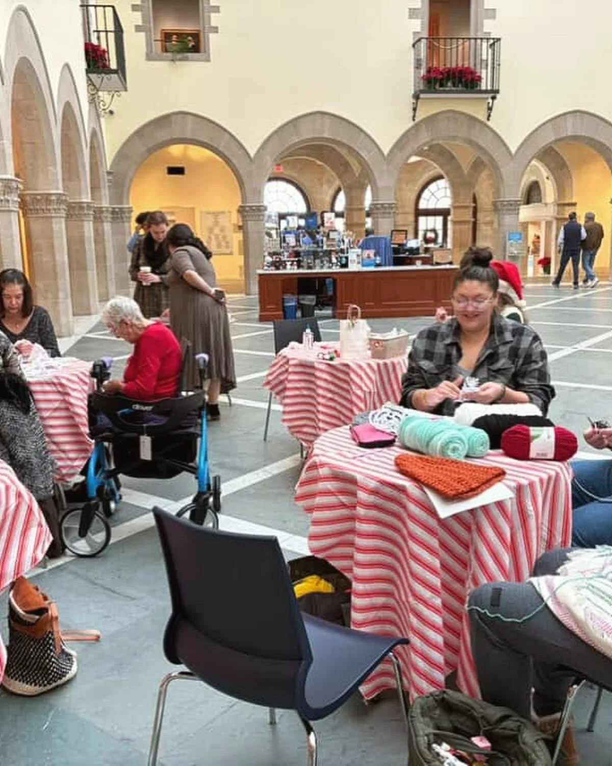 People sitting at tables with red and white striped tablecloths, knitting and talking inside a large, arch-covered indoor space with a high ceiling and stone arches.
