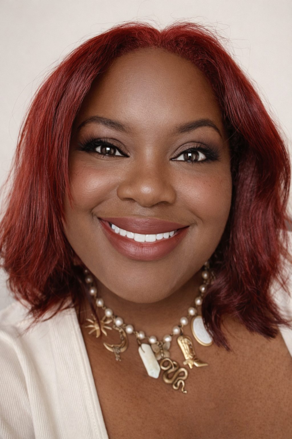 Close-up of a woman with red hair smiling, wearing makeup, a pearl necklace with charms, and a white top.