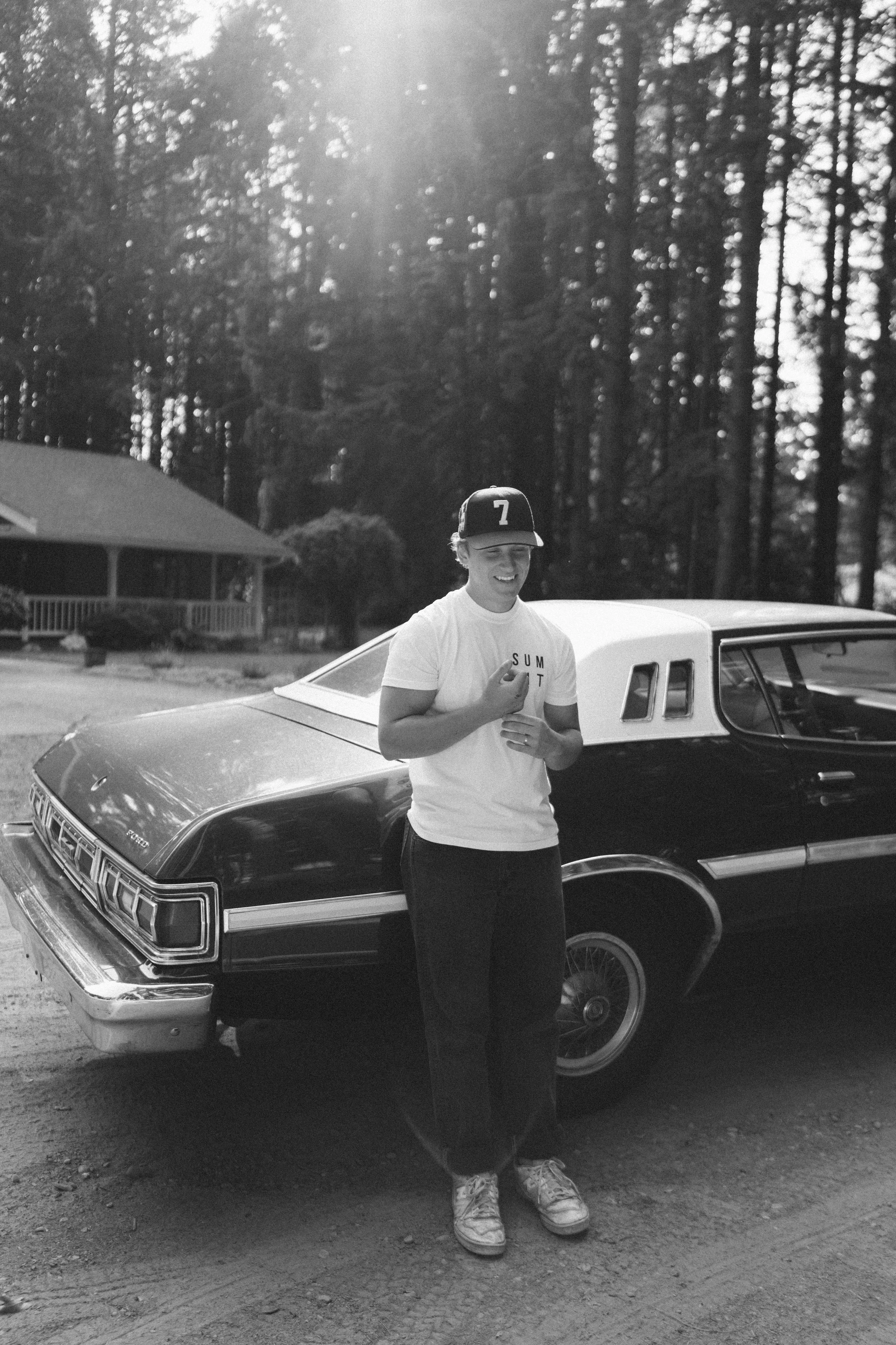 A young person smiling and standing next to a vintage black and white car in a wooded area, wearing a black baseball cap, a white t-shirt, and sneakers, with sunlight filtering through the trees.
