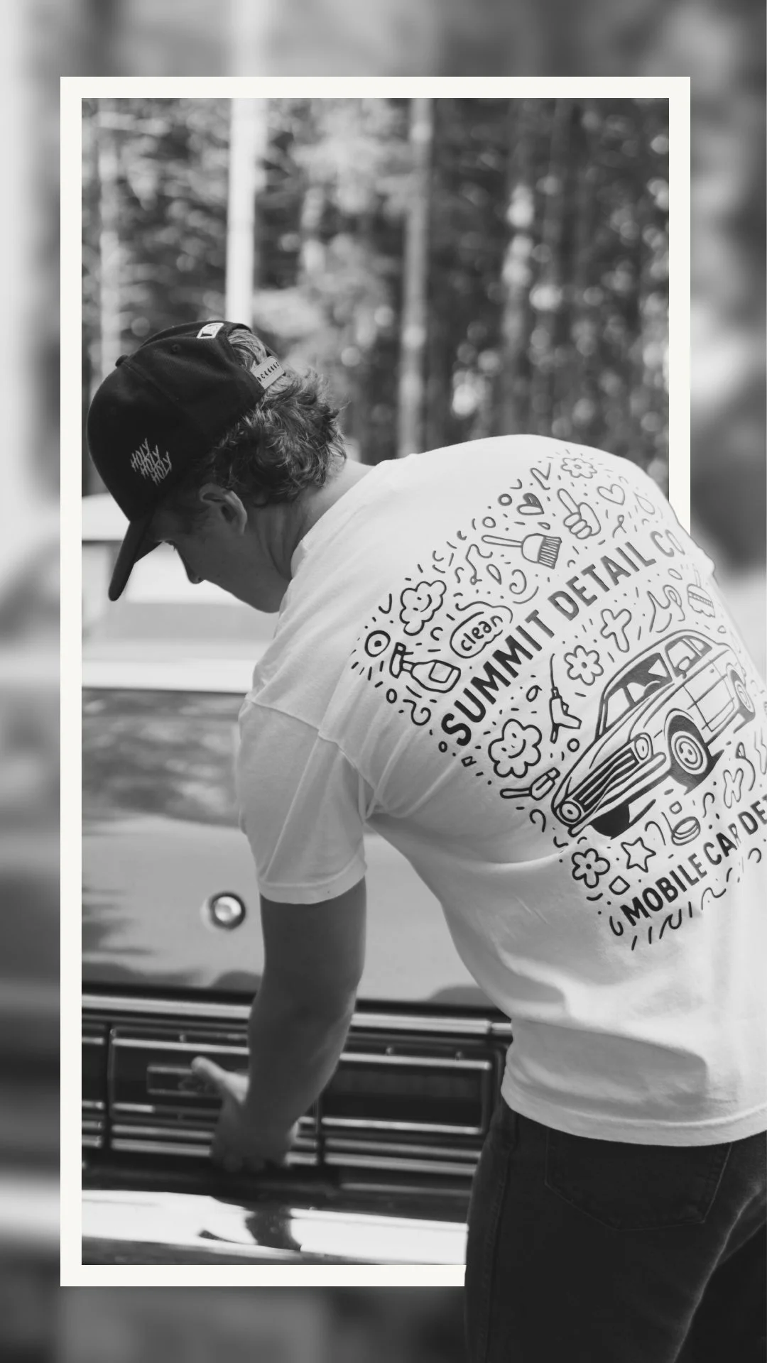 A person wearing a hat and a t-shirt with the words "SUMMIT DETAIL" and a car illustration, cleaning or detailing a vehicle outdoors. The image is in black and white.