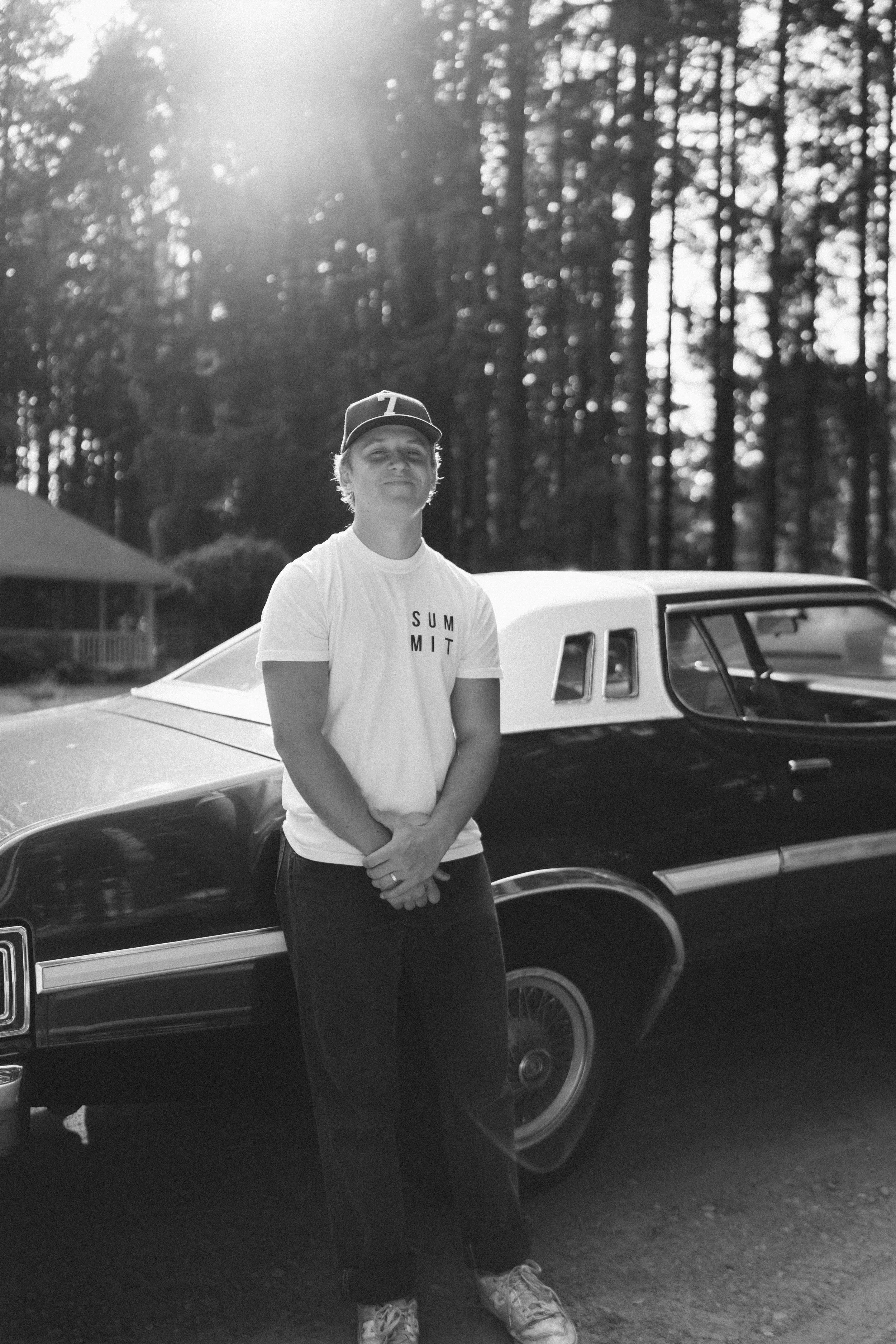 A young man standing in front of a classic car outdoors during the daytime, with trees and sunlight in the background. He is wearing a cap, a white t-shirt with the word 'SUMMIT' printed on it, and dark pants.
