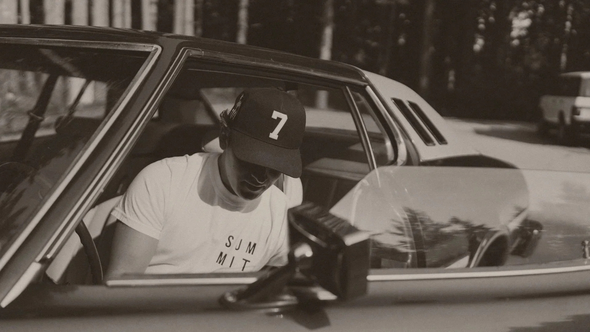 A young man in sunglasses and a cap with the number 7, sitting inside a vintage car with a forested background.