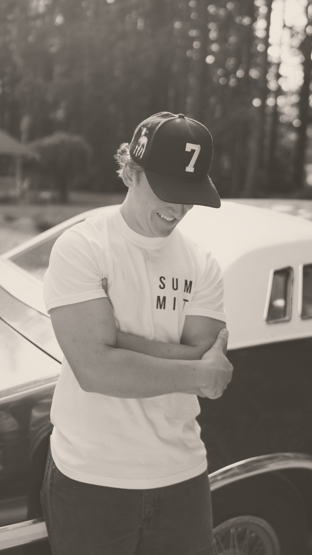 A young man wearing a cap with the number 7 and a white t-shirt with the word 'SUMMIT' on it, smiling and giving a thumbs-up, standing next to a vintage car outdoors in a wooded area.