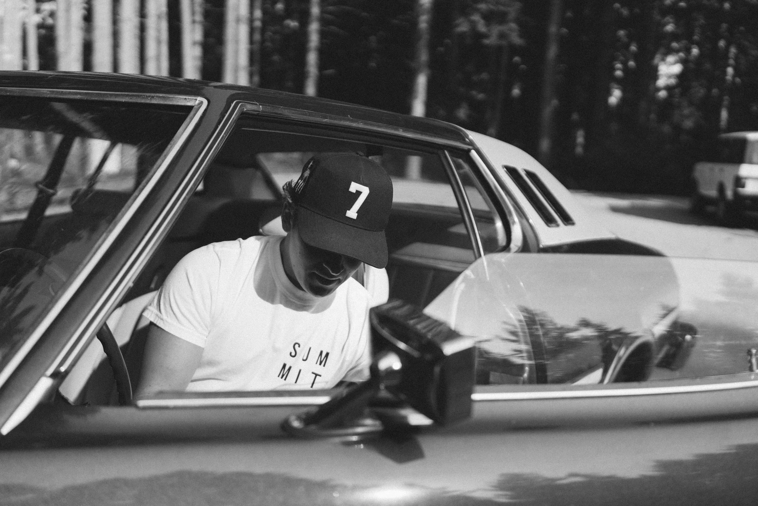 A young man in a baseball cap and sunglasses sitting in the driver seat of a vintage car, looking downward, with trees and another car visible outside.
