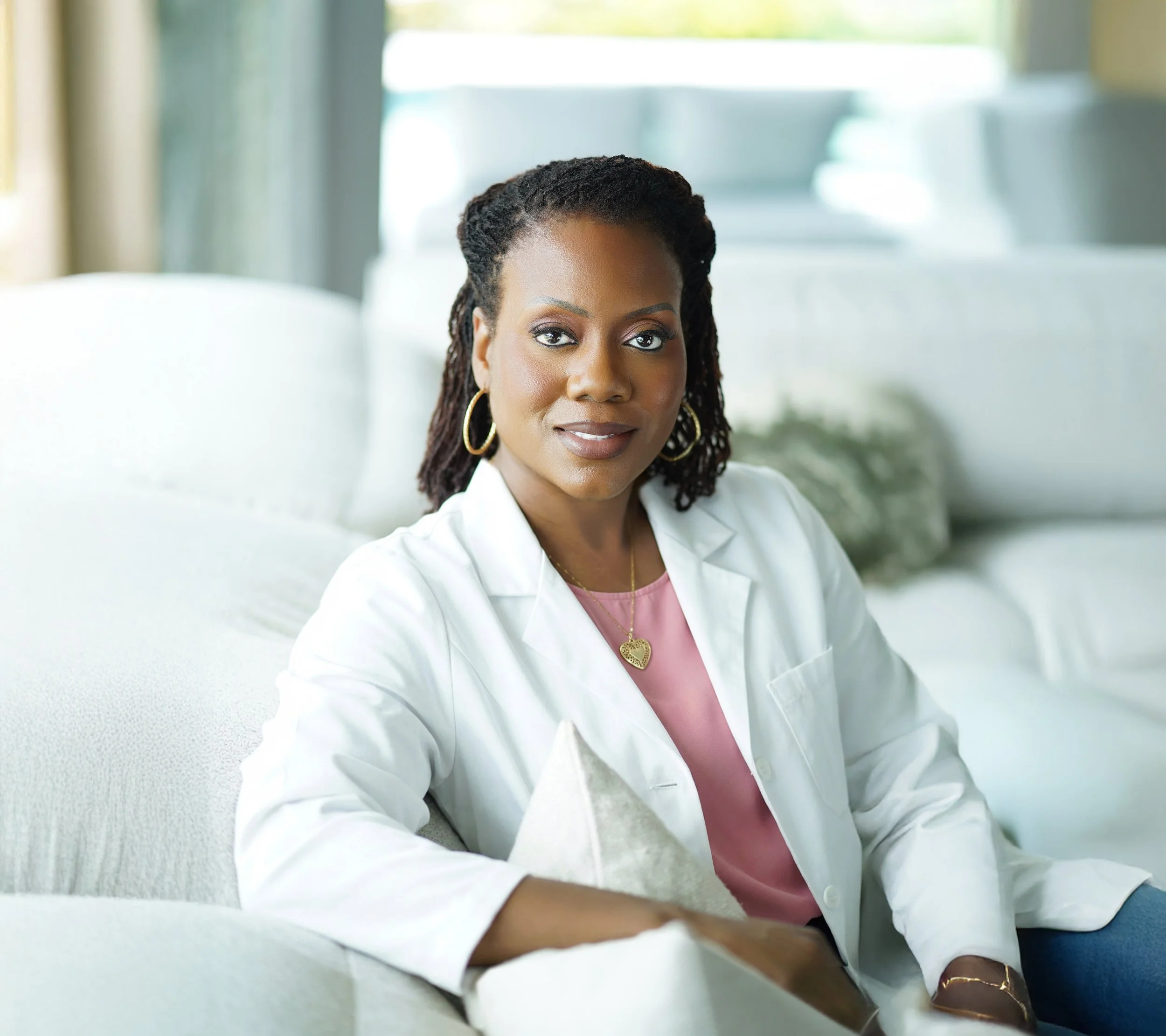 Dr Jessica White Videa a white blazer and pink top sitting on a light-colored sofa in a bright, modern living room.