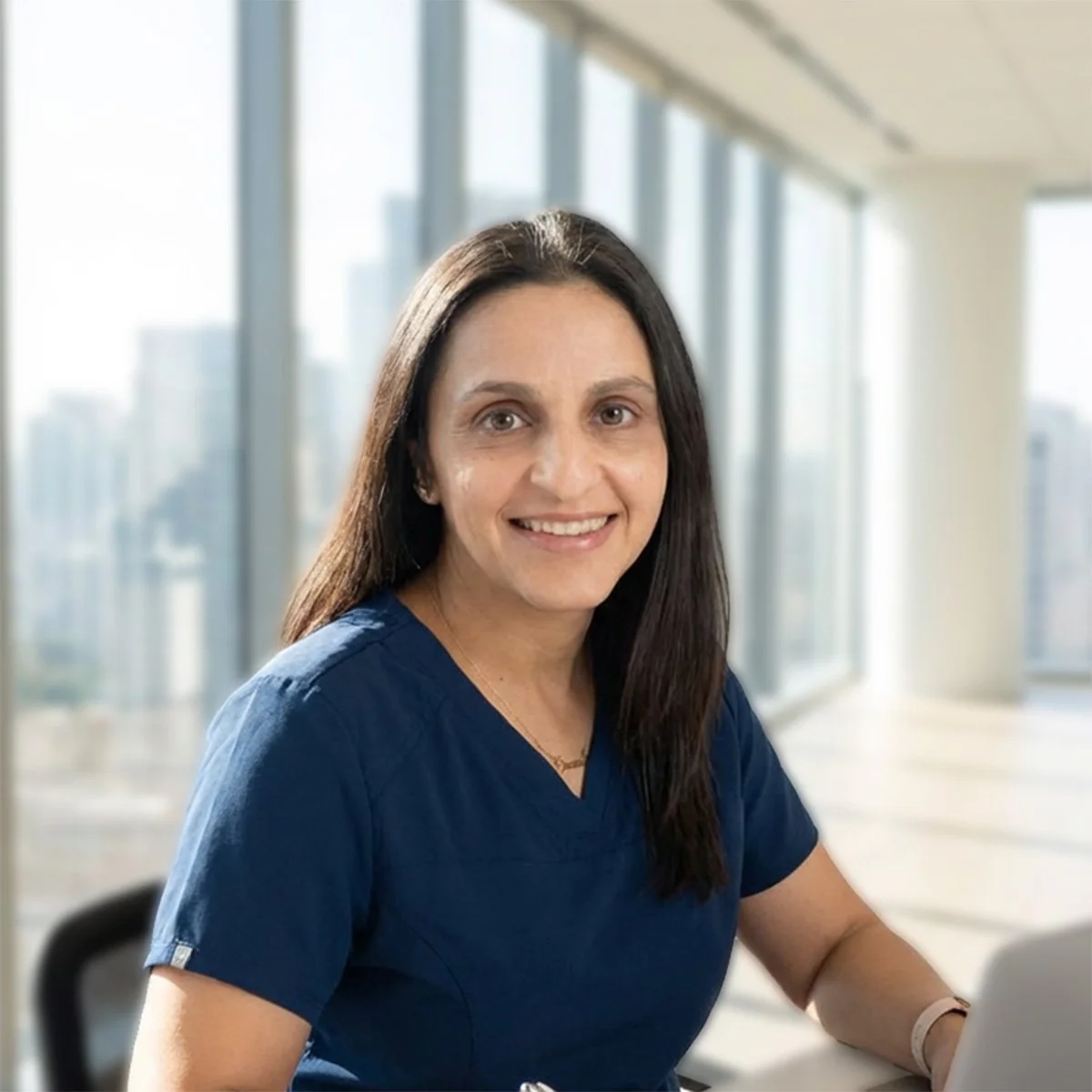 A woman wearing blue scrubs sitting at a desk in an office with large windows and a city view, smiling at the camera.