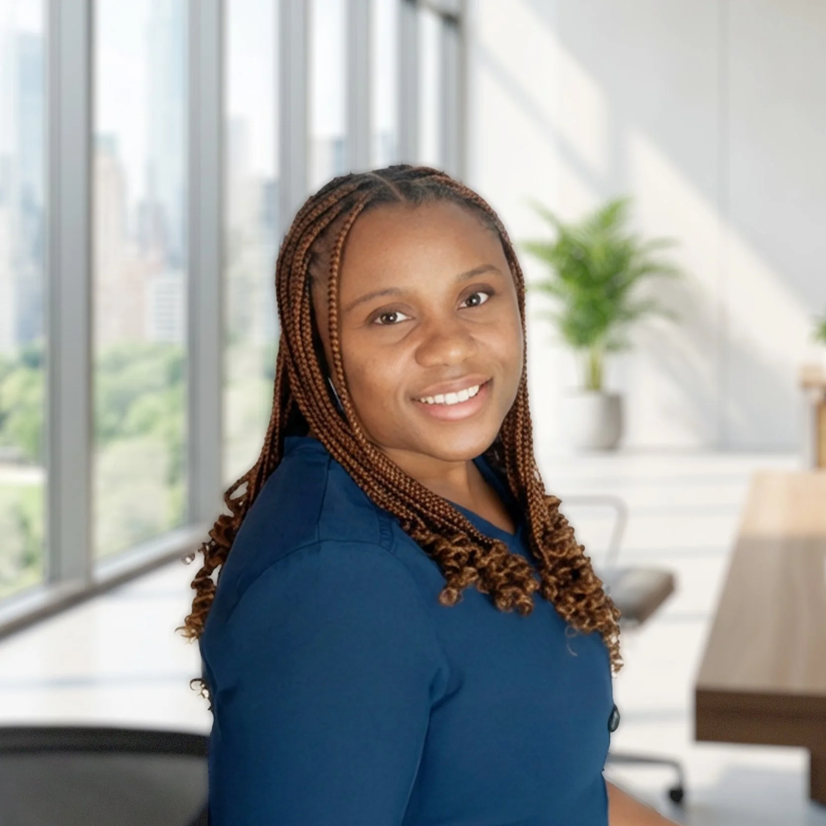 A young woman with braids wearing a blue shirt, smiling, in an office with large windows and a potted plant in the background.