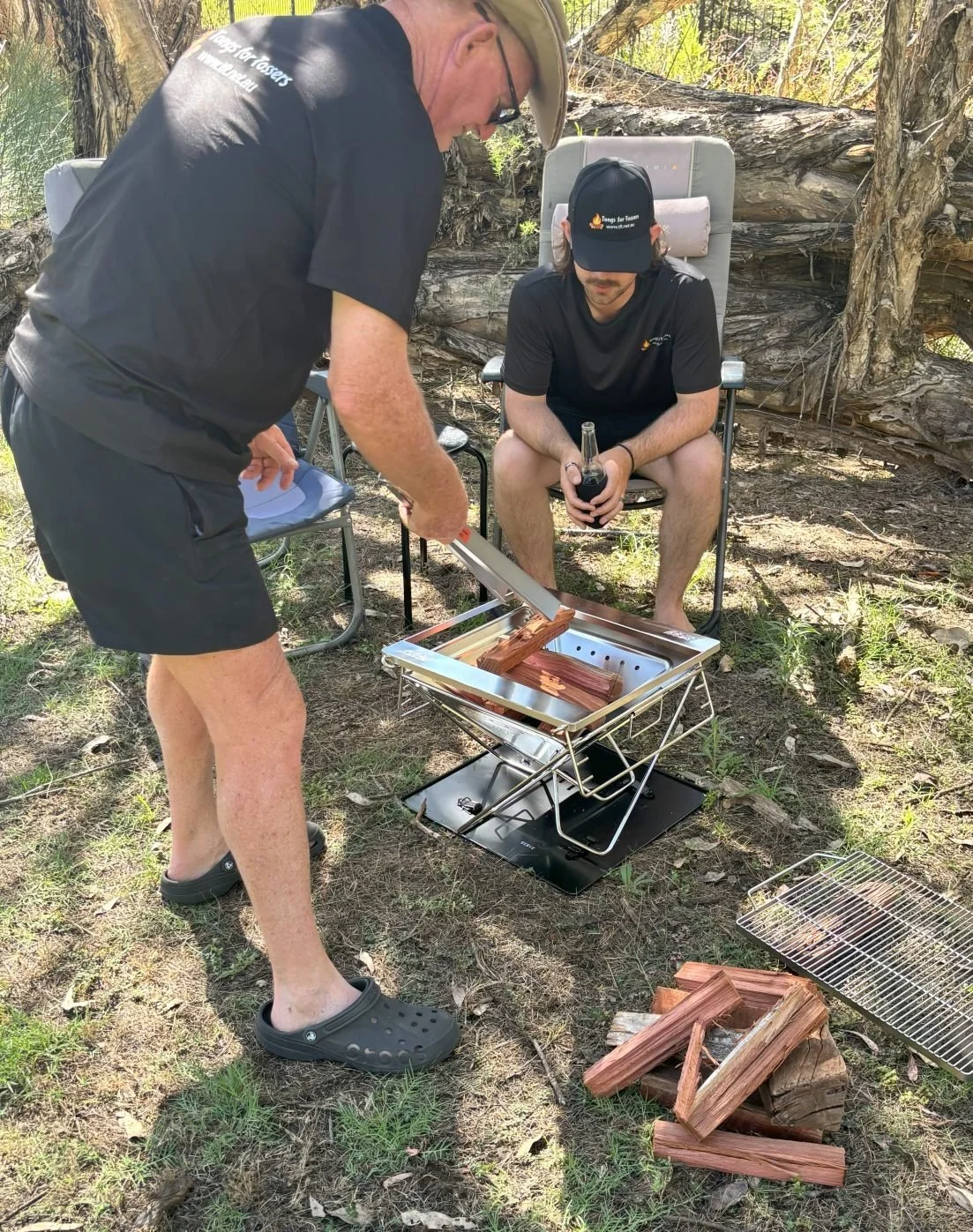 Two men outdoors with a set of Tongs for Tossers with a fire pit between them.