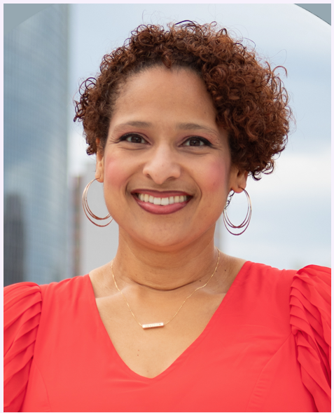 A woman with curly reddish-brown hair smiling outdoors, wearing a red top, hoop earrings, and a gold necklace with a horizontal bar pendant.