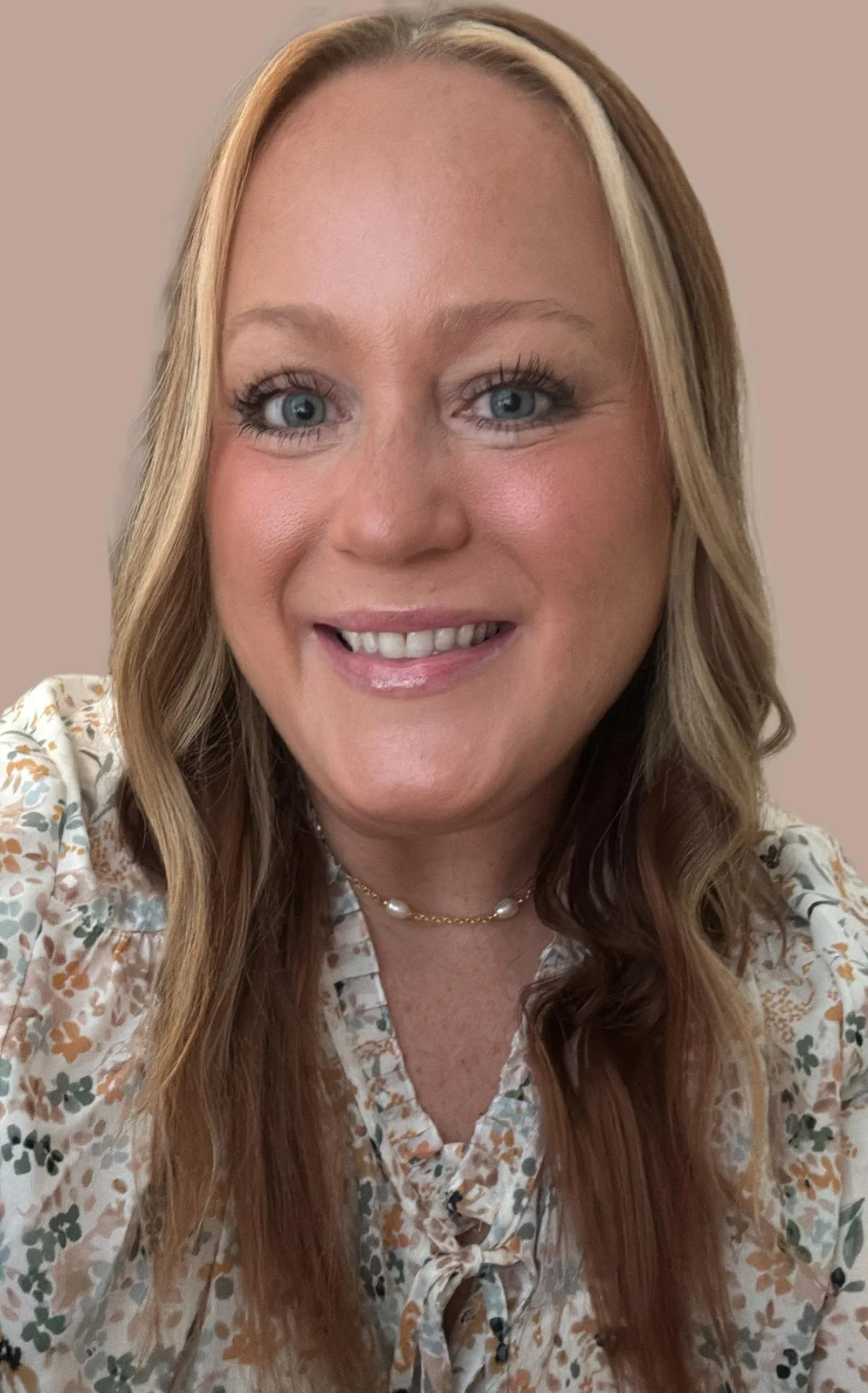 A woman with long wavy hair, blue eyes, and a bright smile taking a selfie. She is wearing a floral blouse and a pearl necklace.