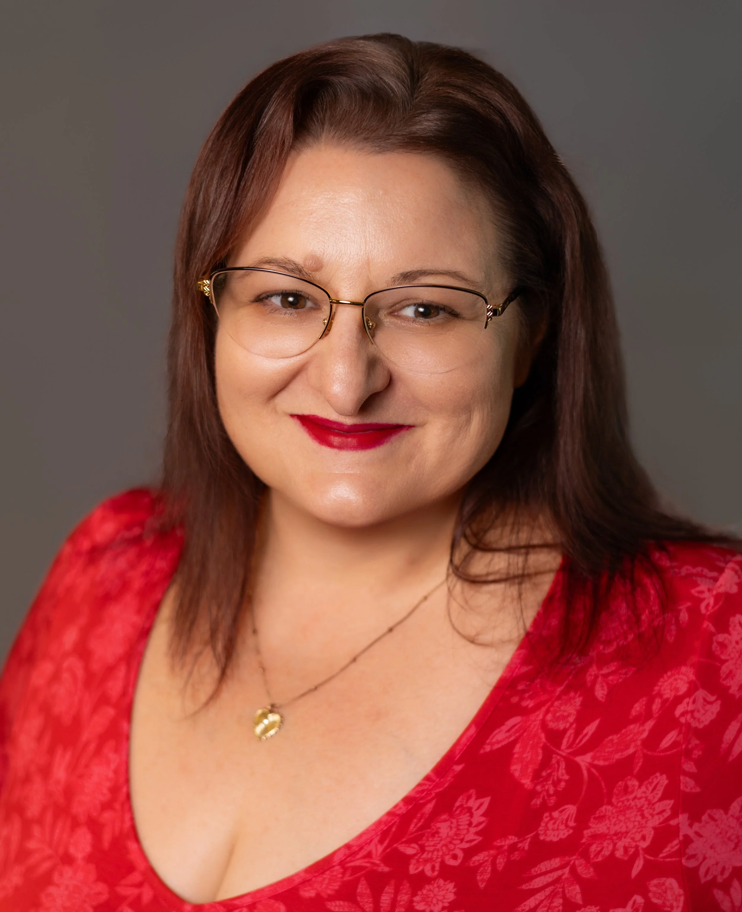 A woman with brown hair, wearing glasses, red lipstick, and a red floral top. She has a necklace with a gold pendant and is smiling at the camera against a neutral gray background.