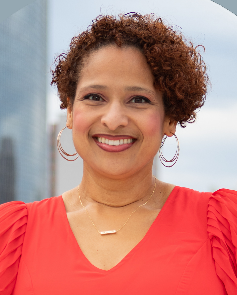 A woman with short curly hair smiling, wearing hoop earrings, a gold necklace, and a red top, outdoors with modern buildings in the background.