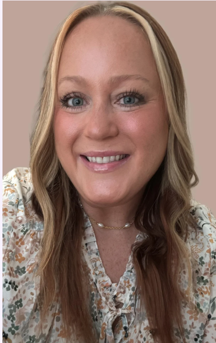 Smiling woman with long blonde hair, blue eyes, wearing a floral blouse and a necklace, posing against a plain background.