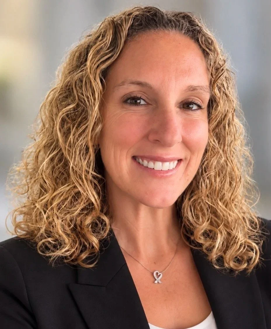 A woman with shoulder-length curly blonde hair, smiling, wearing a black blazer and a necklace with a heart-shaped pendant.