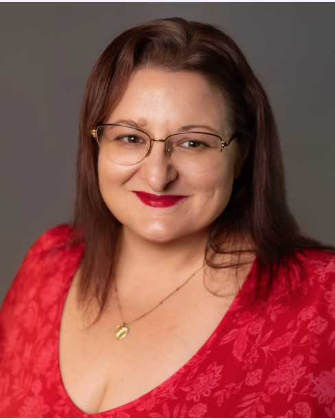 Close-up of a woman with shoulder-length brown hair, wearing glasses, a red floral blouse, and red lipstick, smiling at the camera against a gray background.
