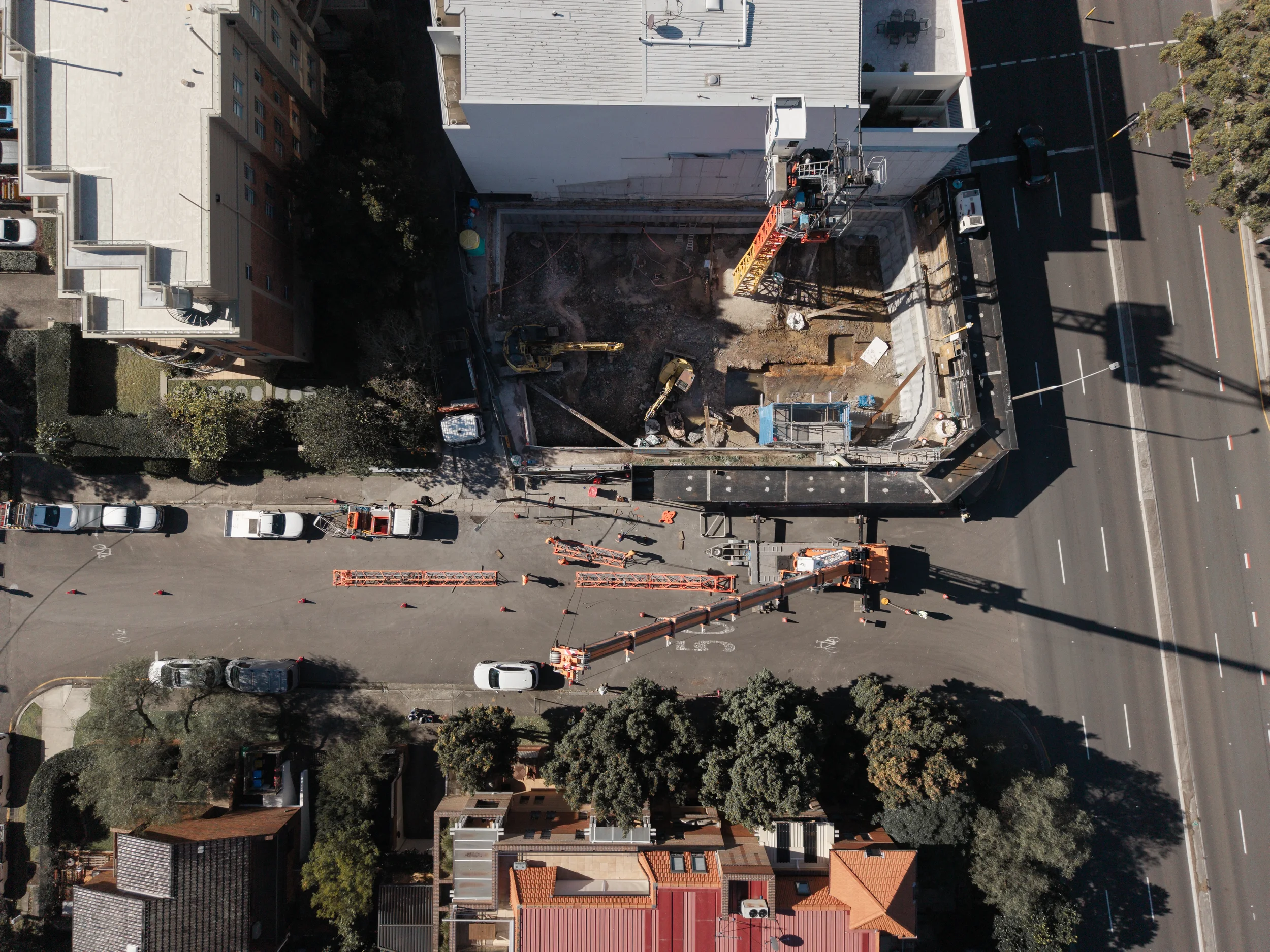 An aerial view of a construction site in an urban area with a partially excavated area surrounded by a fence, construction equipment, and a crane, with nearby buildings and streets.