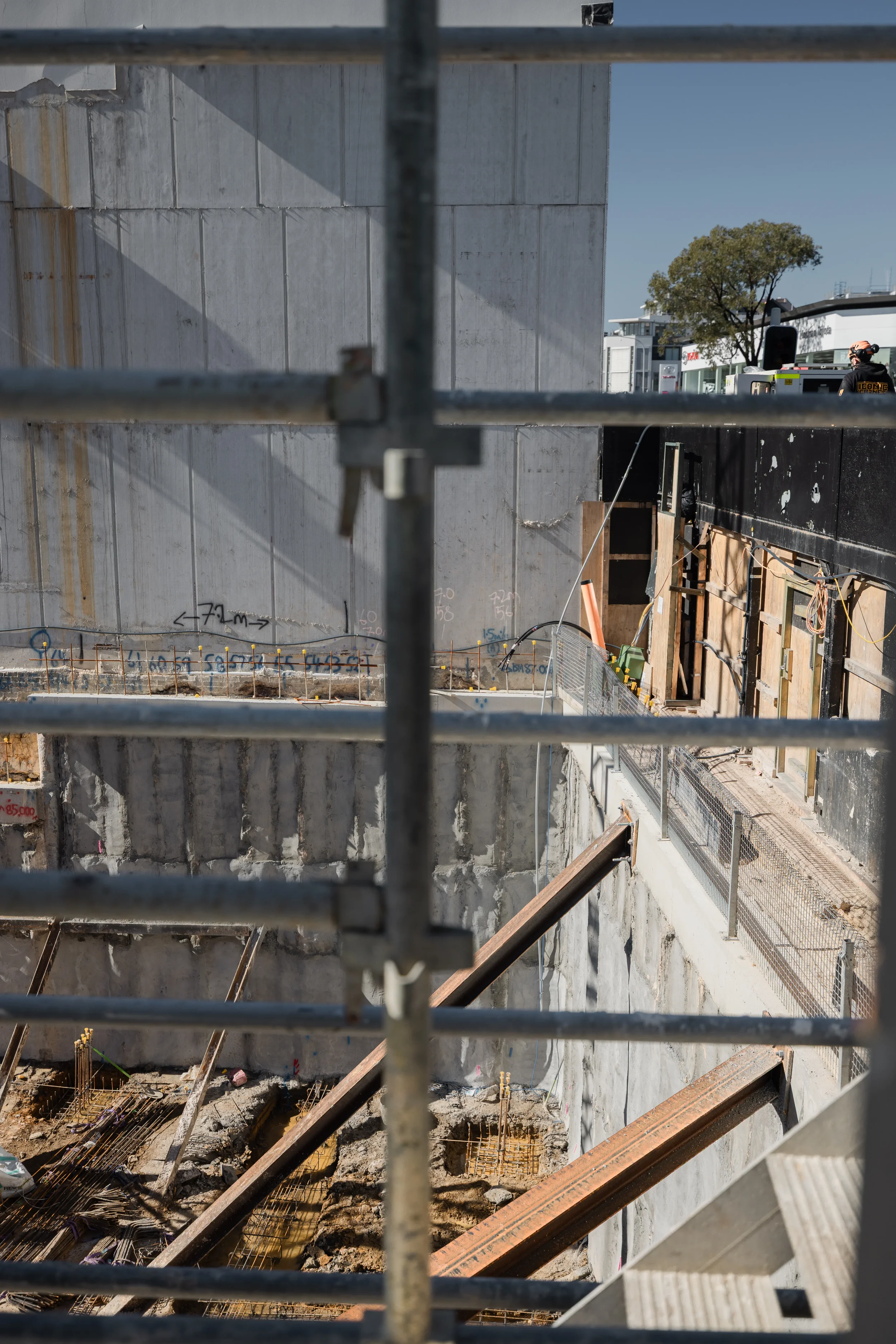 Construction site viewed through metal scaffolding, showing concrete walls, wooden supports, and rebar for ongoing building foundation work in daytime.