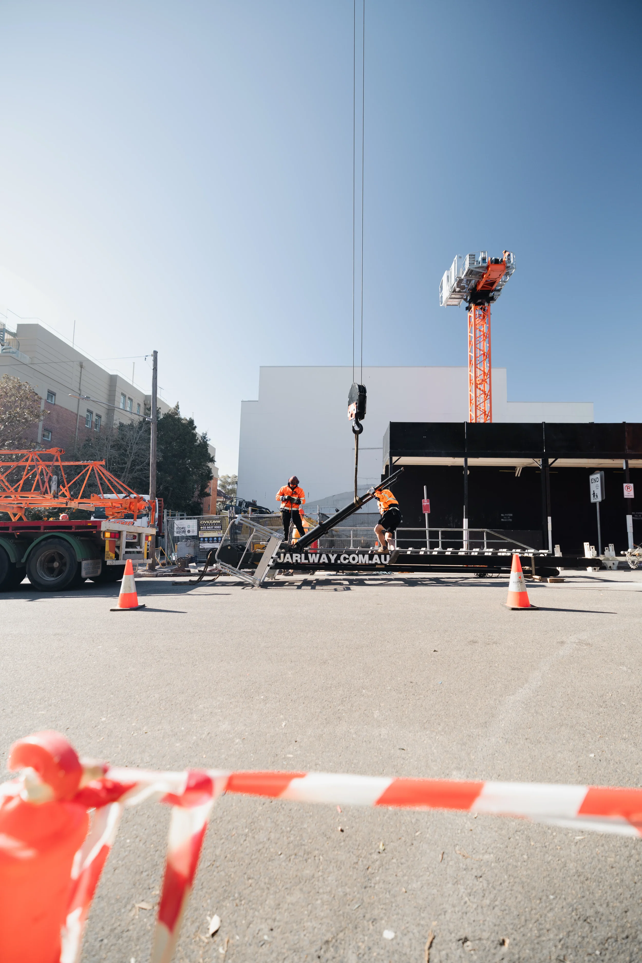 Construction workers setting up equipment on a city street, with a tall construction crane in the background and orange safety cones and barrier tape in the foreground.