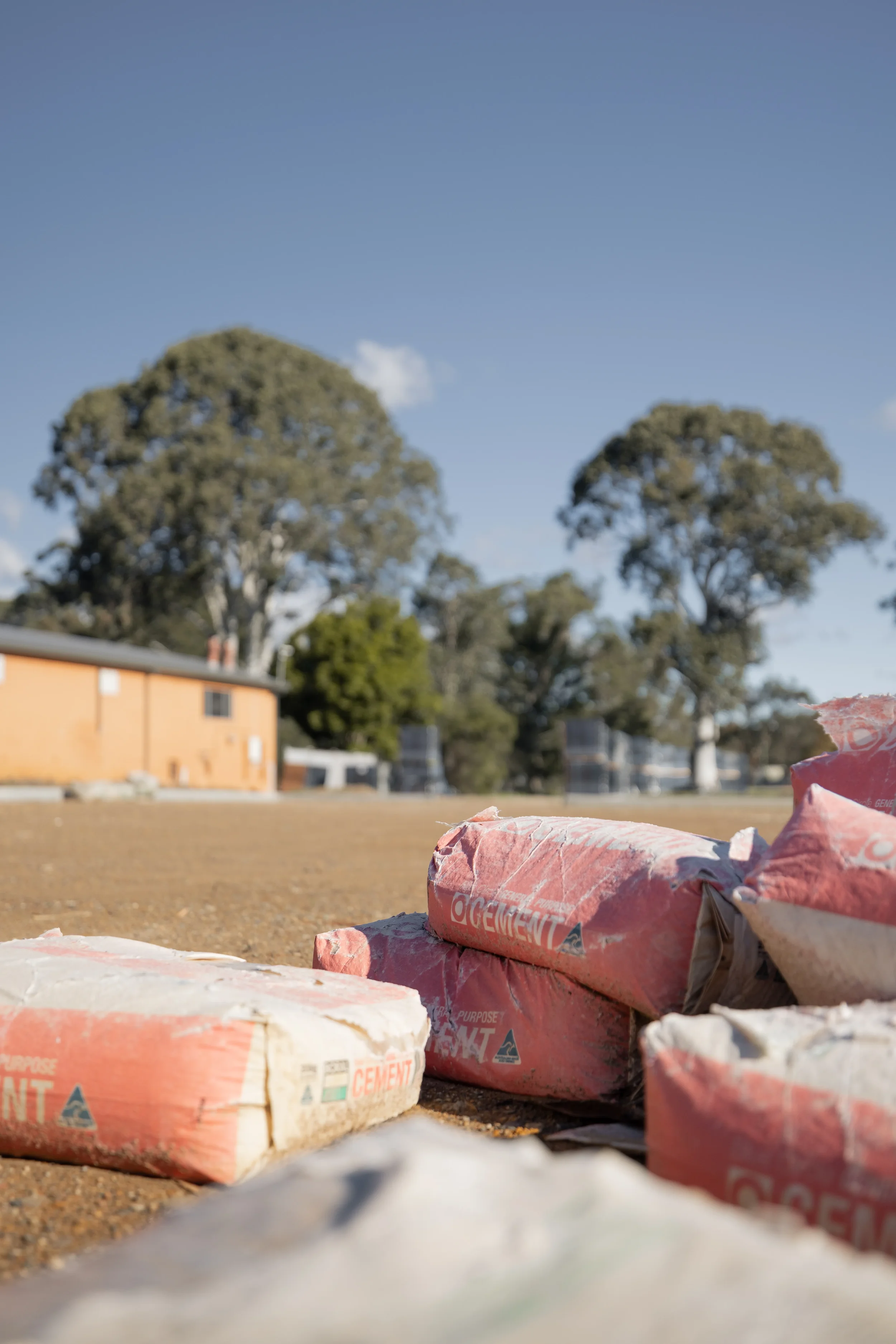 Several bags of cement stacked on the ground outdoors, with a building and trees in the background and a clear blue sky.