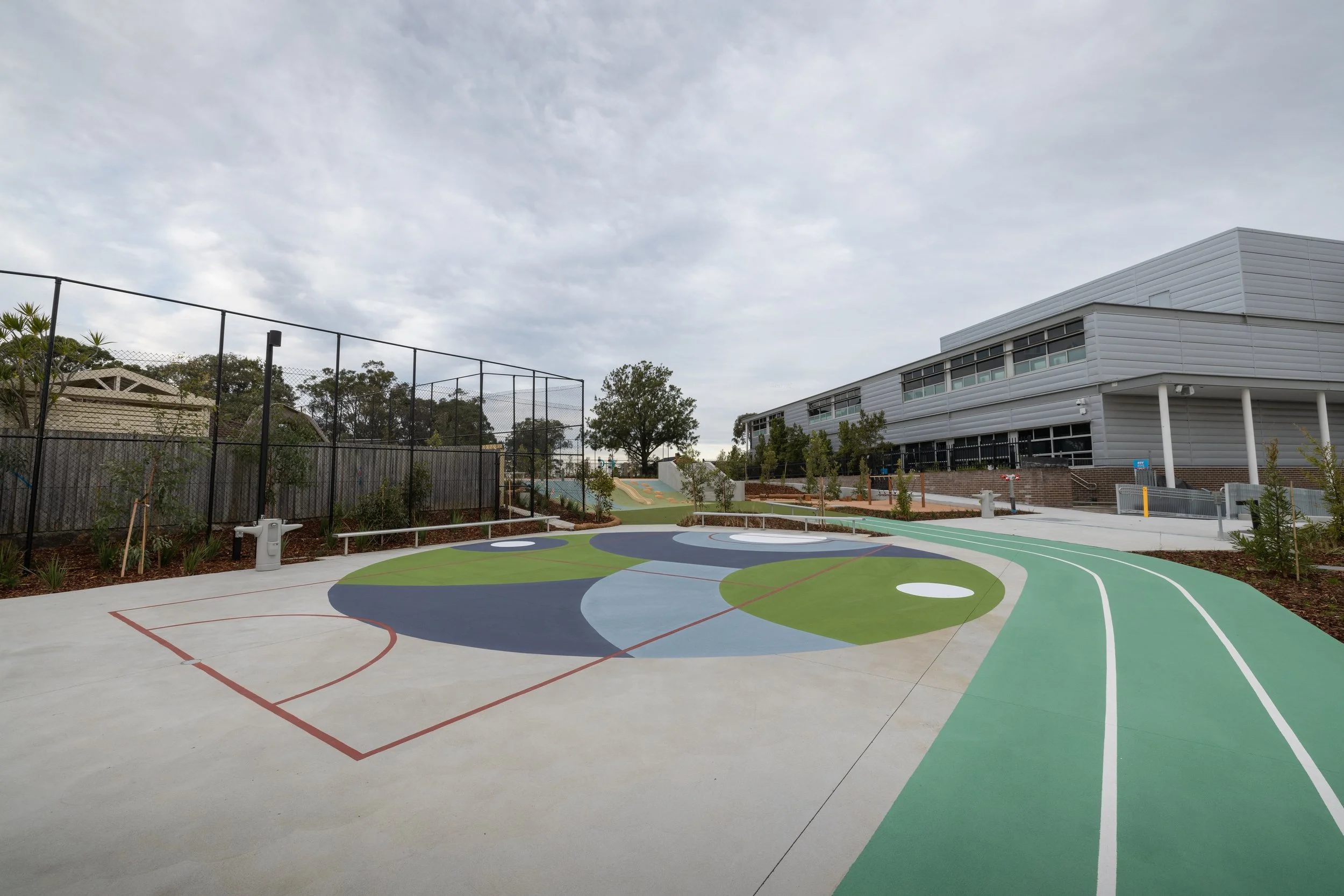 Outdoor playground with colorful abstract sports court, a fenced area, trees, and a modern building under a cloudy sky.