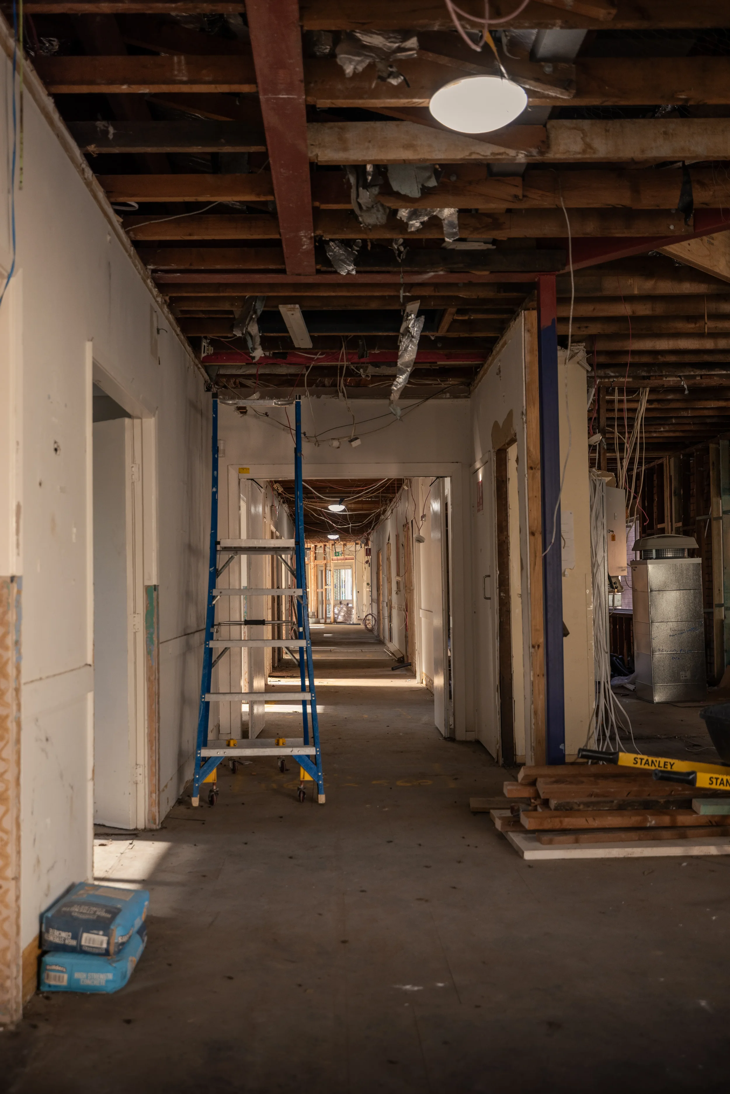 A hallway under renovation with exposed wooden ceiling beams, wiring, and construction materials. A blue step ladder is in the center, and stacks of drywall or insulation are on the floor to the left. The hallway leads into multiple rooms, with some 