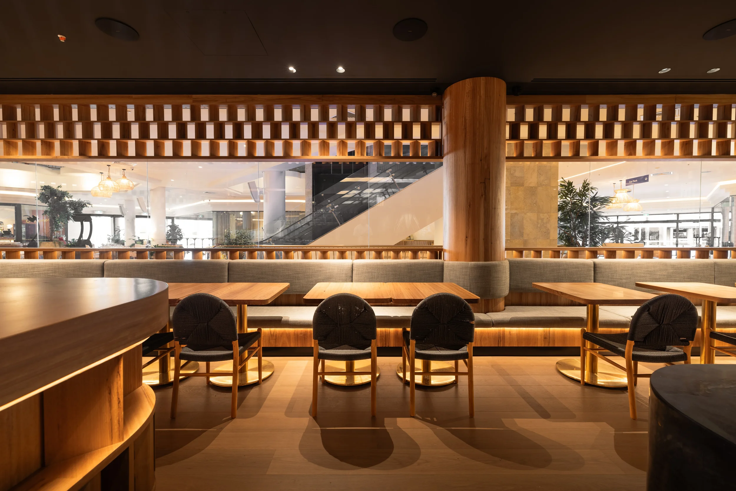 Interior of a modern restaurant or café with wooden furniture, including tables and chairs, and a decorative wooden grid partition. There are large glass windows, plants, and warm lighting fixtures.