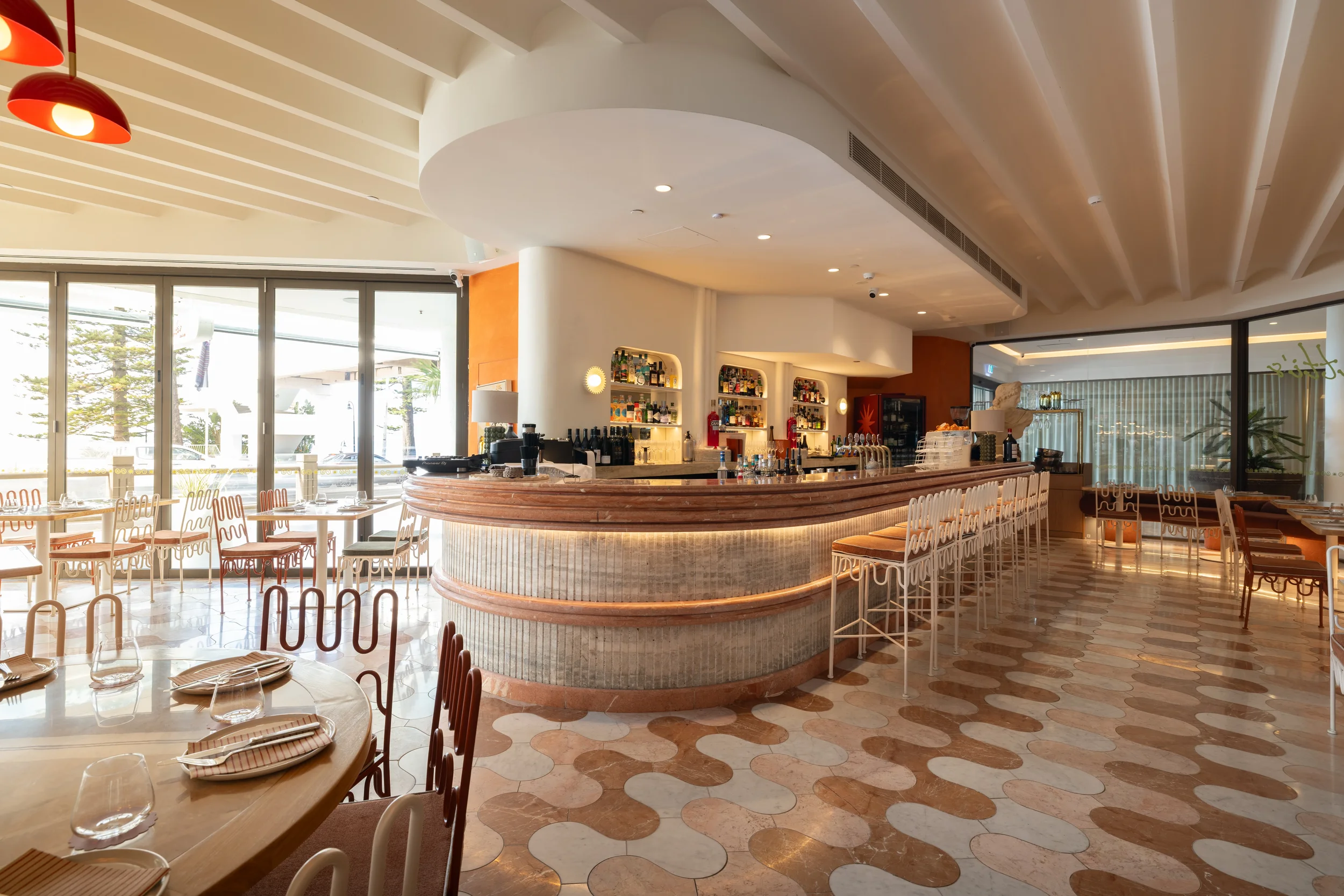 Interior of a modern restaurant with a curved bar, seating area, tables set with glasses and napkins, large windows letting in natural light, and a ceiling with white beams.