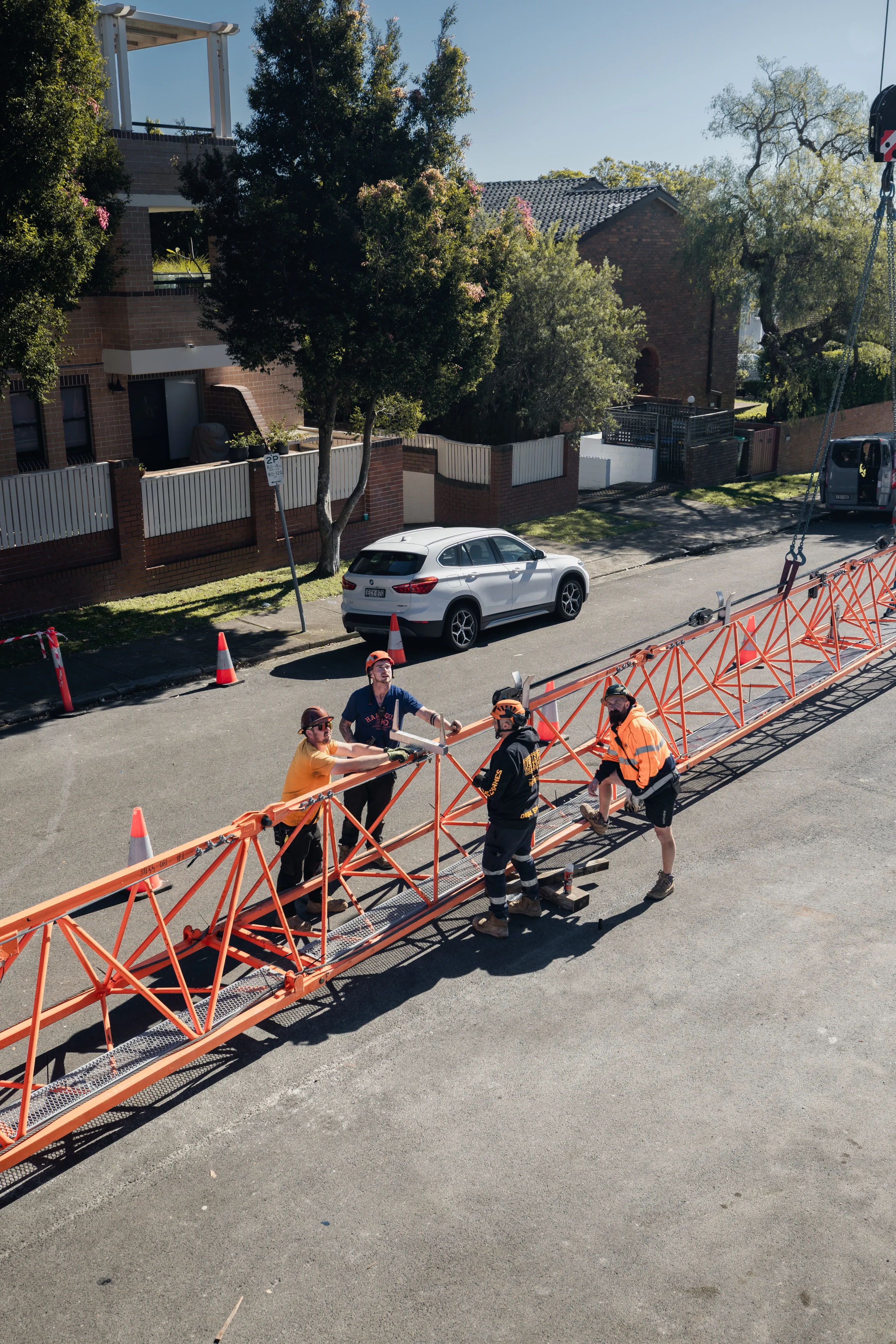 Construction workers wearing helmets and safety gear assembling orange scaffolding on a residential street with parked cars, trees, and houses in the background.