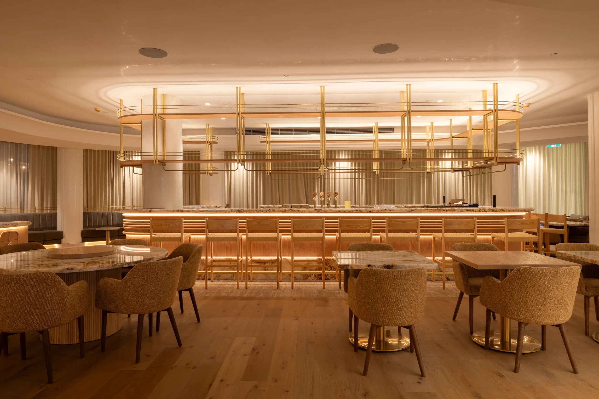 Interior of a hotel lounge or bar with wooden chairs and tables, a marble counter, and a large modern gold-colored chandelier hanging from the ceiling, with curtains in the background.