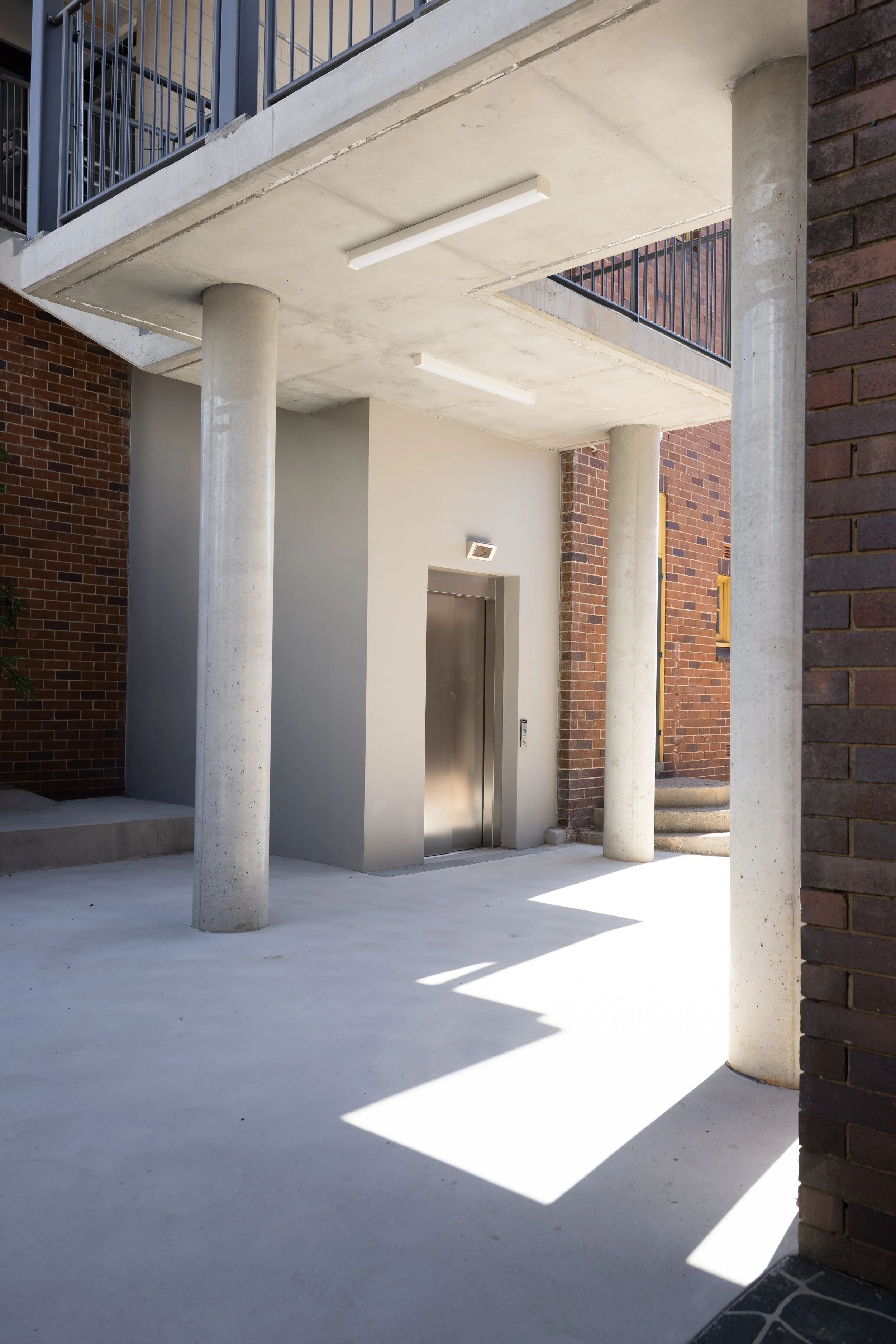 Modern building entrance with concrete columns, an elevator door, brick walls, and shadows on the ground.