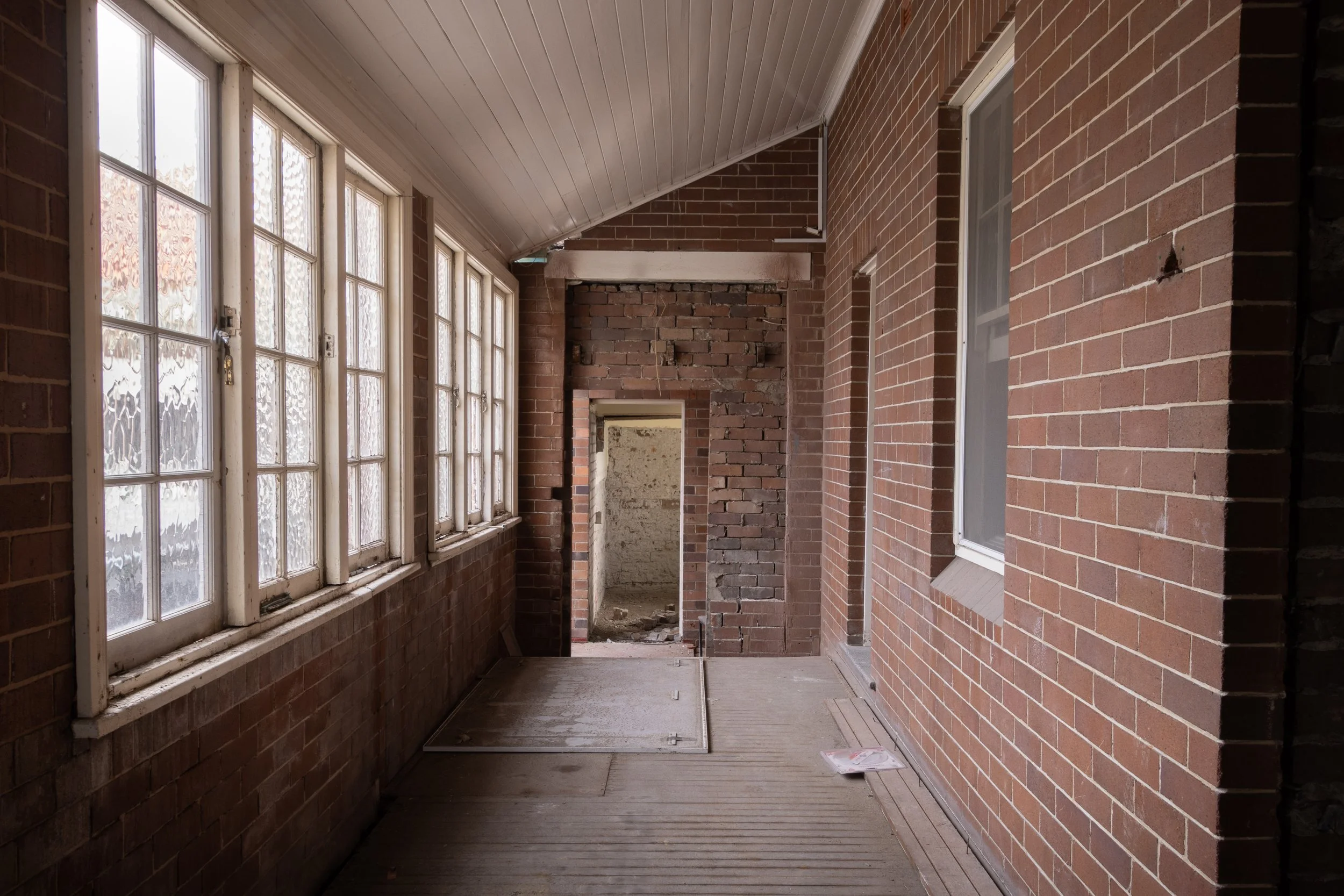 A covered porch or walkway with brick walls, wooden flooring, and large windows with multiple panes.