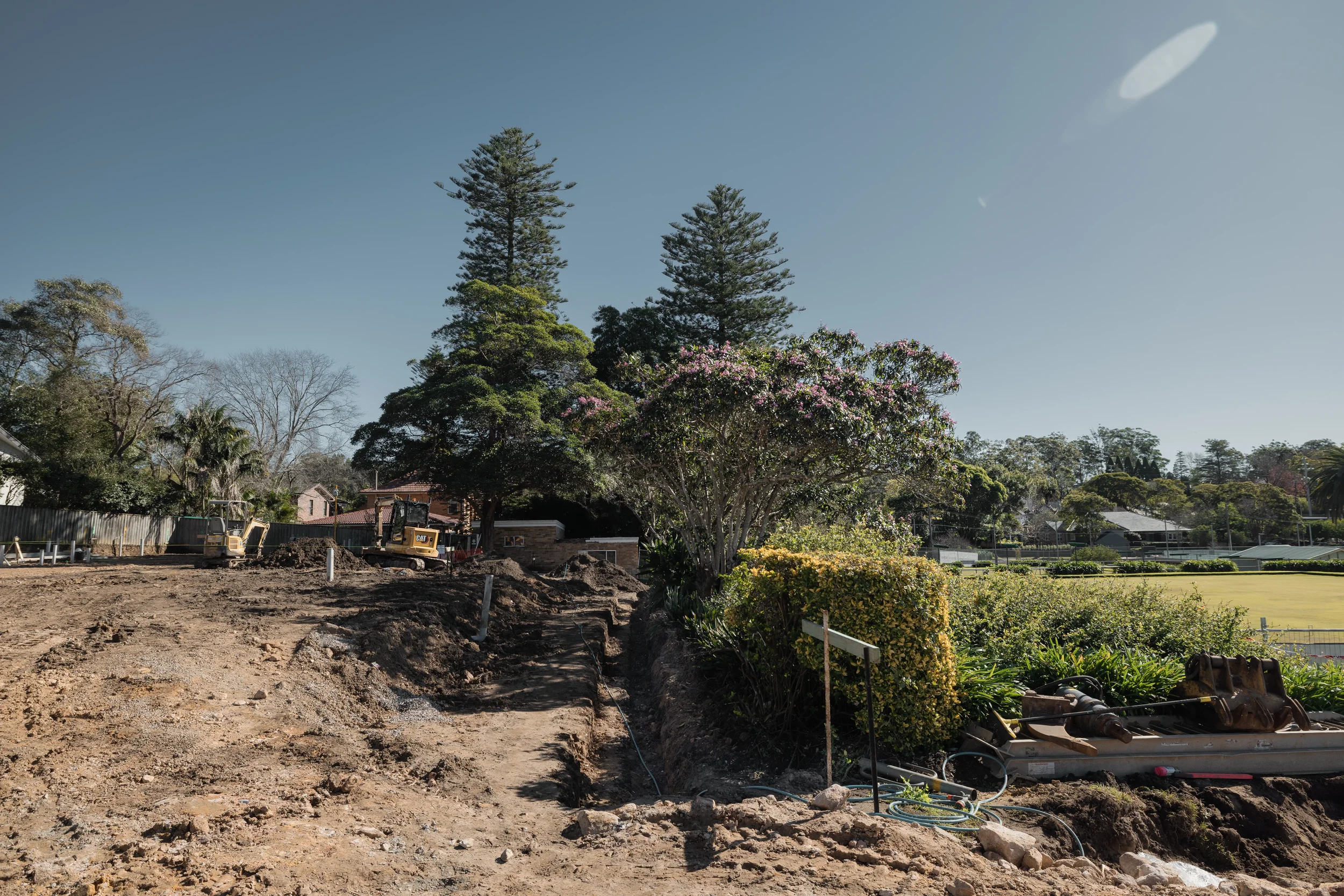 Construction site with dirt and soil, construction equipment, and green trees under a clear blue sky.