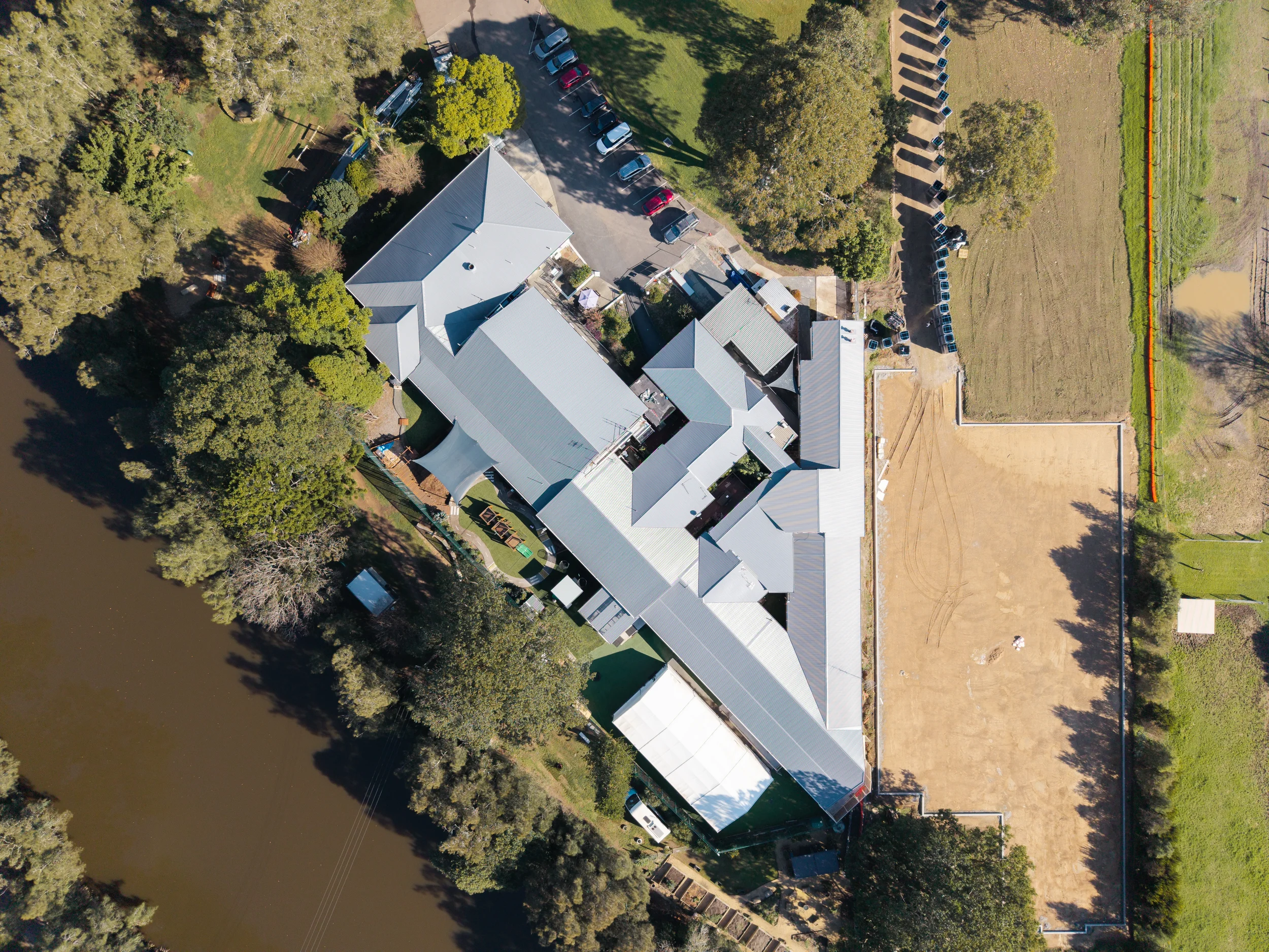 Aerial view of a property with multiple buildings, trees, a parking lot with cars, and a nearby sports field with tennis courts surrounded by trees and a water body.