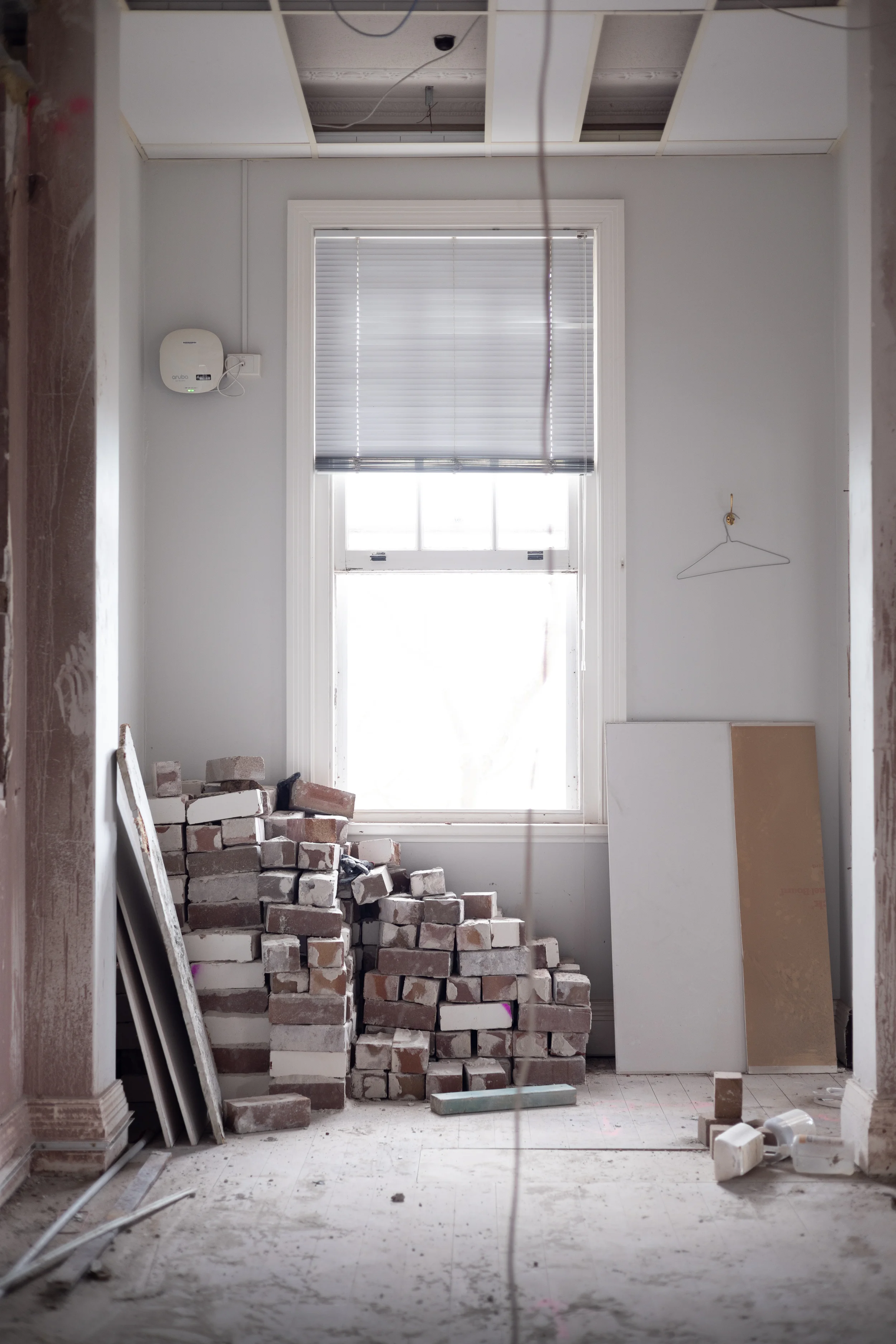 Interior room under construction with a tall window, a pile of bricks in the center, construction materials and tools scattered on the floor, and an unfinished ceiling with exposed wiring.
