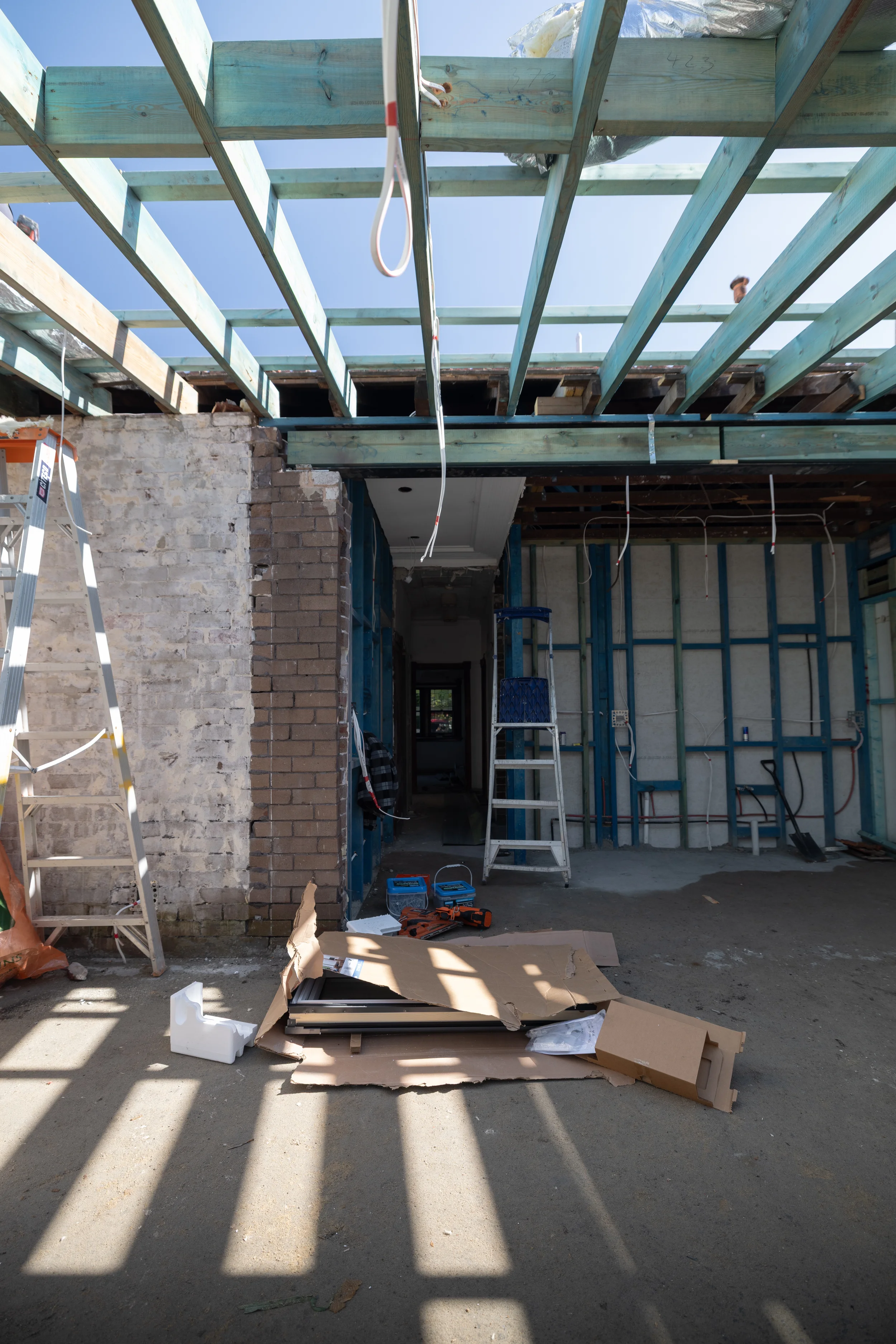 Construction site with framing for a building, several ladders, tools, and scattered materials including cardboard boxes and construction supplies, under a clear blue sky.