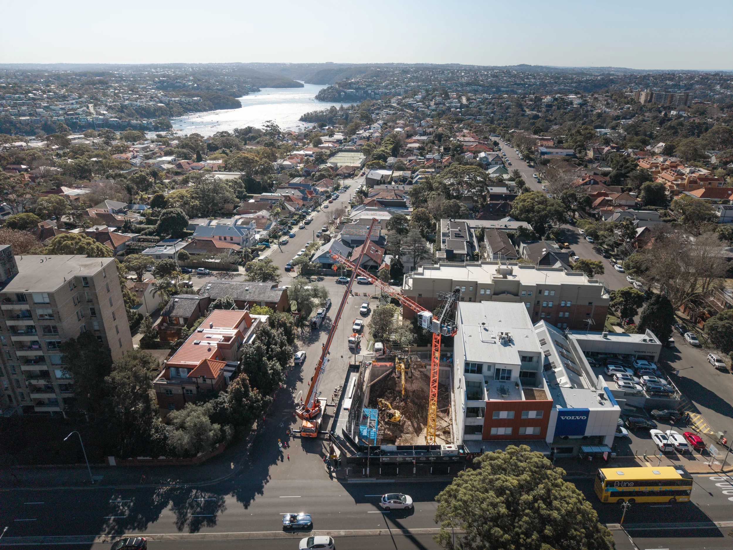 An aerial view of a city with a river, residential houses, and construction site with cranes, adjacent to a multi-lane road with vehicles including a yellow double-decker bus.